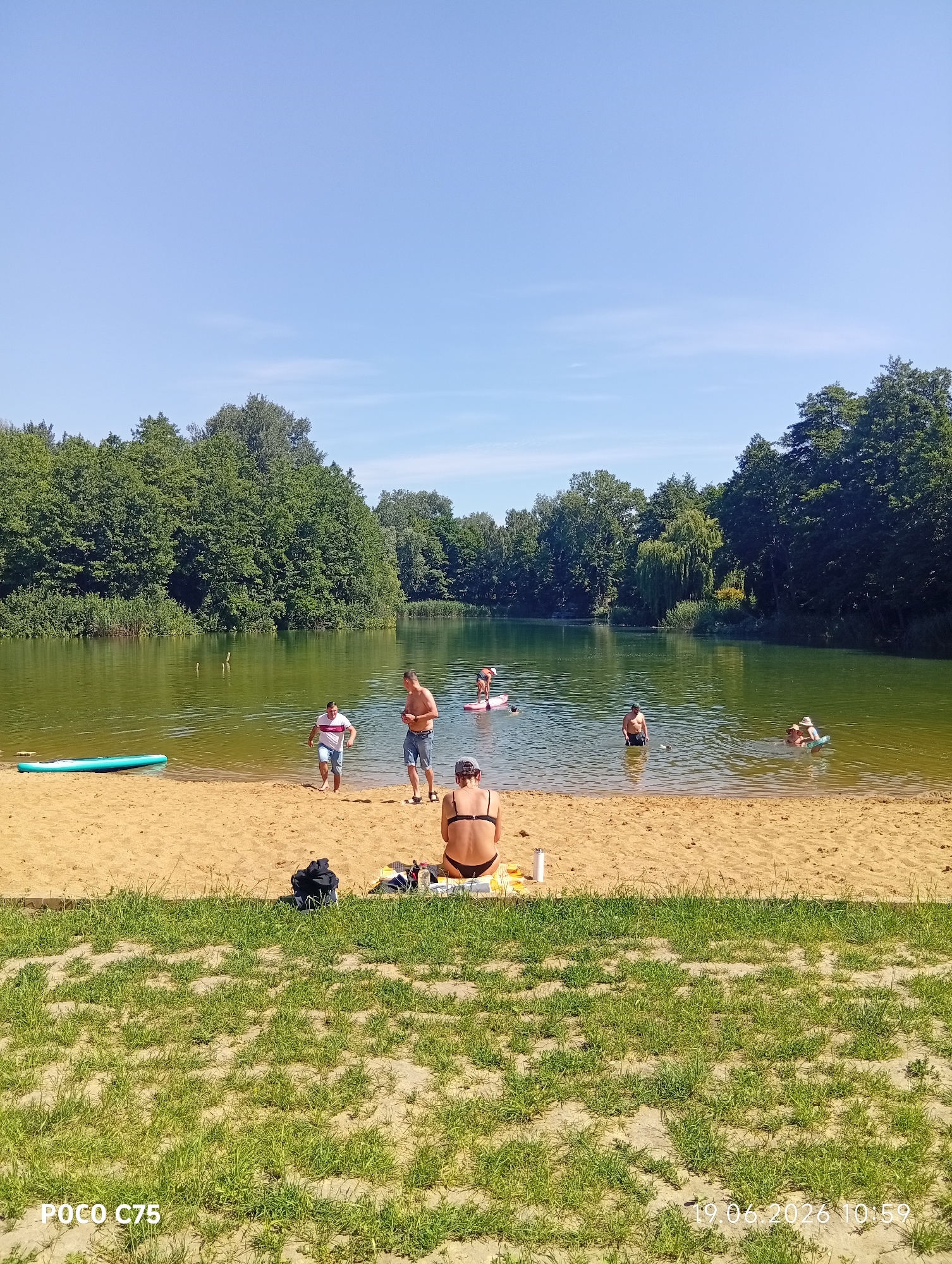 This is a high-angle, full shot of a sandy beach and a lake on a sunny day. The beach area in the foreground is covered with sand and grass. Several people are scattered across the scene, some on the beach and others in the water.
On the beach, a woman in a black bikini sits facing away from the camera, her back to the viewer. To her left, a black backpack and other items are placed on the sand. Further back, two men are near the water's edge. One man, wearing a white t-shirt and shorts, is walking towards the water, and another man in denim shorts is standing and looking towards the water. There is a light blue paddleboard on the sand near the water.
In the lake, several people are engaged in various water activities. Two people are on a paddleboard in the mid-ground. Further out in the water, other individuals can be seen swimming or wading. The lake is surrounded by lush green trees, forming a dense forest that extends to the horizon. The sky above is clear and blue, indicating fair weather. The timestamp "19.06.2026 10:59" is visible in the bottom right corner of the image. The watermark "POCO C75" is in the bottom left.
This scene is consistent with a recreational area or park, likely a popular spot for swimming and water sports during warm weather. Given the timestamp format and the presence of such features, this could be a location in or around Wrocław, Poland.