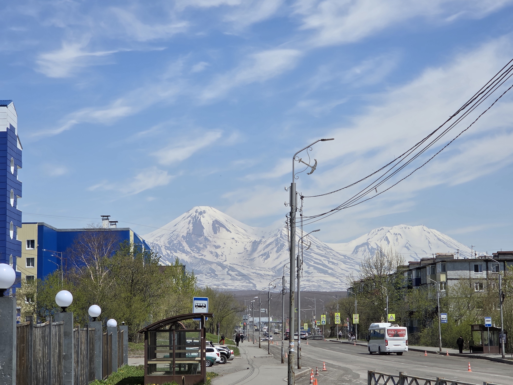 The image captures a broad urban street scene under a bright blue sky with scattered wispy white clouds. Two majestic, snow-capped mountains, likely volcanoes, dominate the distant background, their white peaks stark against the sky.
In the immediate foreground on the left, a blue and white multi-story building with distinctive circular windows is partially visible. A wooden fence with concrete posts, each topped by a spherical white light fixture, extends along the left side of the street. Behind the fence, a small patch of green grass and several parked cars (including white, green, and dark vehicles) are visible next to a bus stop shelter with a dark frame and transparent panels.
The center and right foreground feature a two-lane asphalt road, partially lined with orange traffic cones suggesting an ongoing activity or temporary lane designation. Multiple utility poles carrying numerous black power lines extend diagonally across the upper right quadrant of the image. One prominent streetlight pole, centrally located, is adorned with a decorative anchor-shaped fixture beneath its lamp. A white minibus, with Russian text "ТУРИЗМ" and "АВАРИЯ" visible on its rear, is traveling away from the viewer in the right lane. Further down the street on the right, another identical bus stop shelter is present.
Several individuals are visible on the sidewalks: some walking, others standing near the bus stops. Various street signs, including a blue square sign with a bus icon (indicating a bus stop), a 40 km/h speed limit sign, and yellow triangular pedestrian crossing signs, are distributed along the road.
The midground displays a mix of residential buildings, some with vibrant blue and yellow facades, others with a more muted grayish concrete finish. Deciduous trees with fresh green foliage are interspersed between the buildings and along the roadside, suggesting spring or early summer. The street recedes into the distance, revealing more vehicles and pedestrians, with the volcanic landscape serving as a dramatic backdrop.
Based on the prominent snow-capped volcanoes and the urban setting, the location is characteristic of Petropavlovsk-Kamchatsky, Russia.