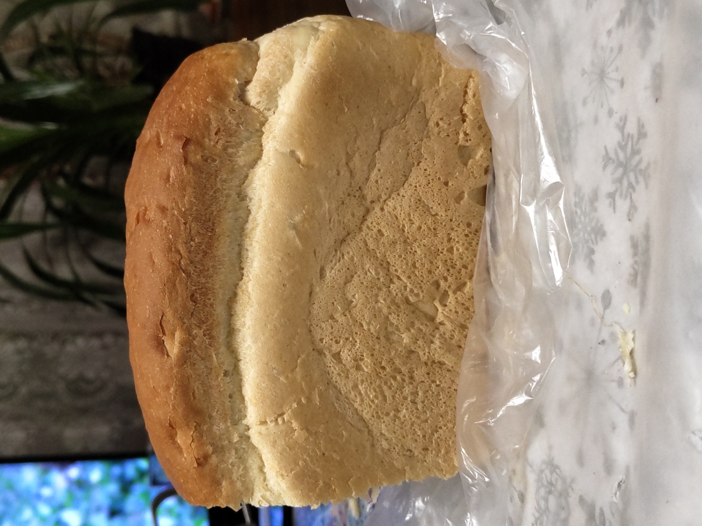 A rectangular loaf of bread, partially cut, rests on crinkled clear plastic film, which is atop a white surface patterned with gray snowflakes. The bread features a golden-brown, somewhat textured crust on its top and a lighter, creamy beige crumb on its exposed vertical face, displaying an irregular, porous internal structure. A small, light-colored crumb or food fragment is visible on the snowflake-patterned surface to the left of the bread. The background is out of focus, showing a bright, blurred blue and white illuminated screen or display on the left, and a dark-colored potted plant with broad leaves on the right, against a dark, possibly patterned wall. The scene is illuminated from the front, highlighting the bread. The location is in the city of Unknown, Unknown.