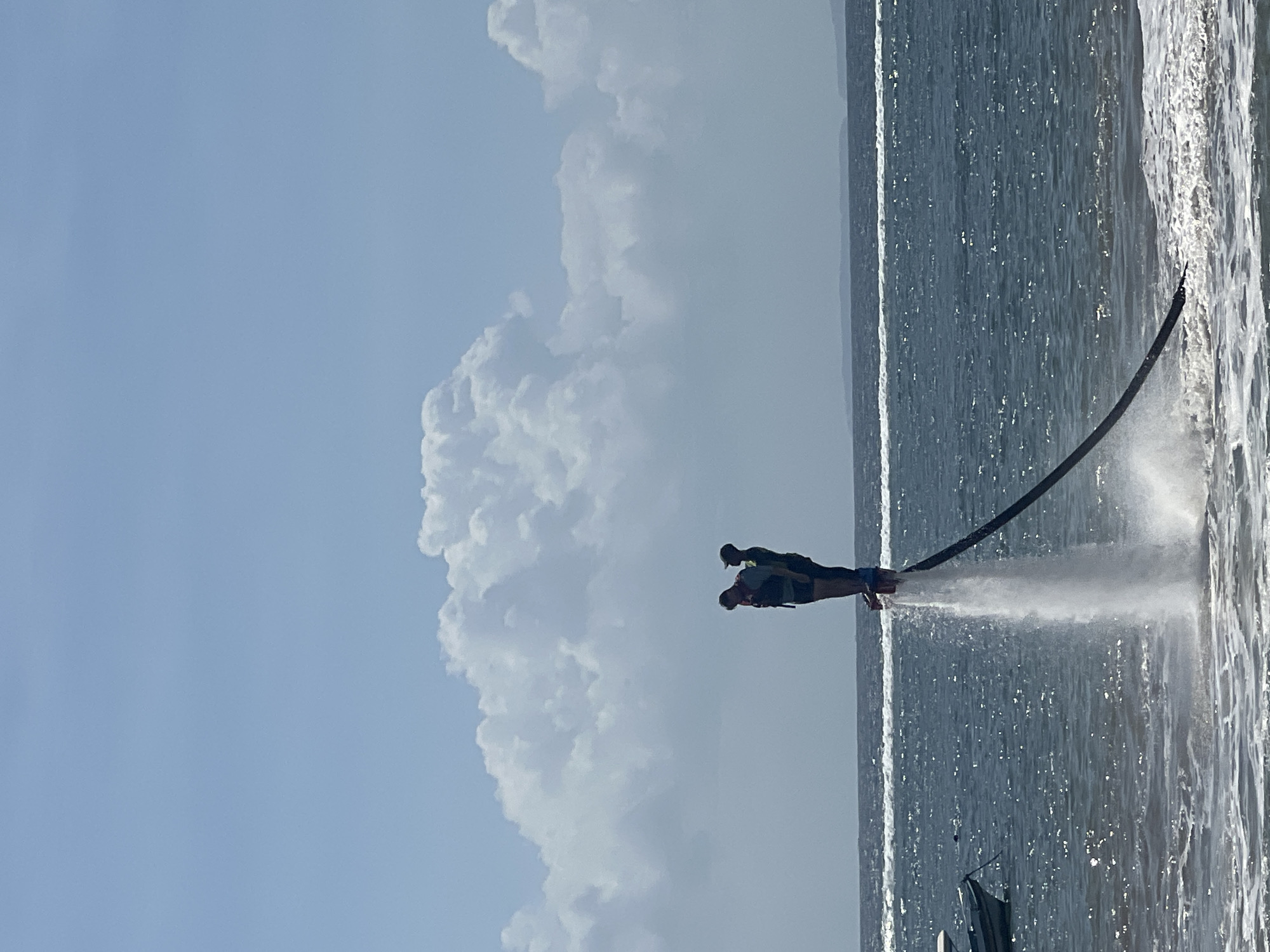 Two individuals are standing on a flyboard, elevated approximately two meters above the ocean surface. The flyboard is propelling water downwards, creating a significant spray and disturbance in the water. The individuals are wearing life vests, with one appearing to be teaching or assisting the other. They are positioned in the ocean, with a body of water extending to the horizon. In the background, there is a coastline with land visible. The sky is blue with scattered white clouds. A small boat is docked to the left of the frame. The overall scene depicts a recreational water sport activity. The location is identified as Kuta Selatan, Indonesia.
