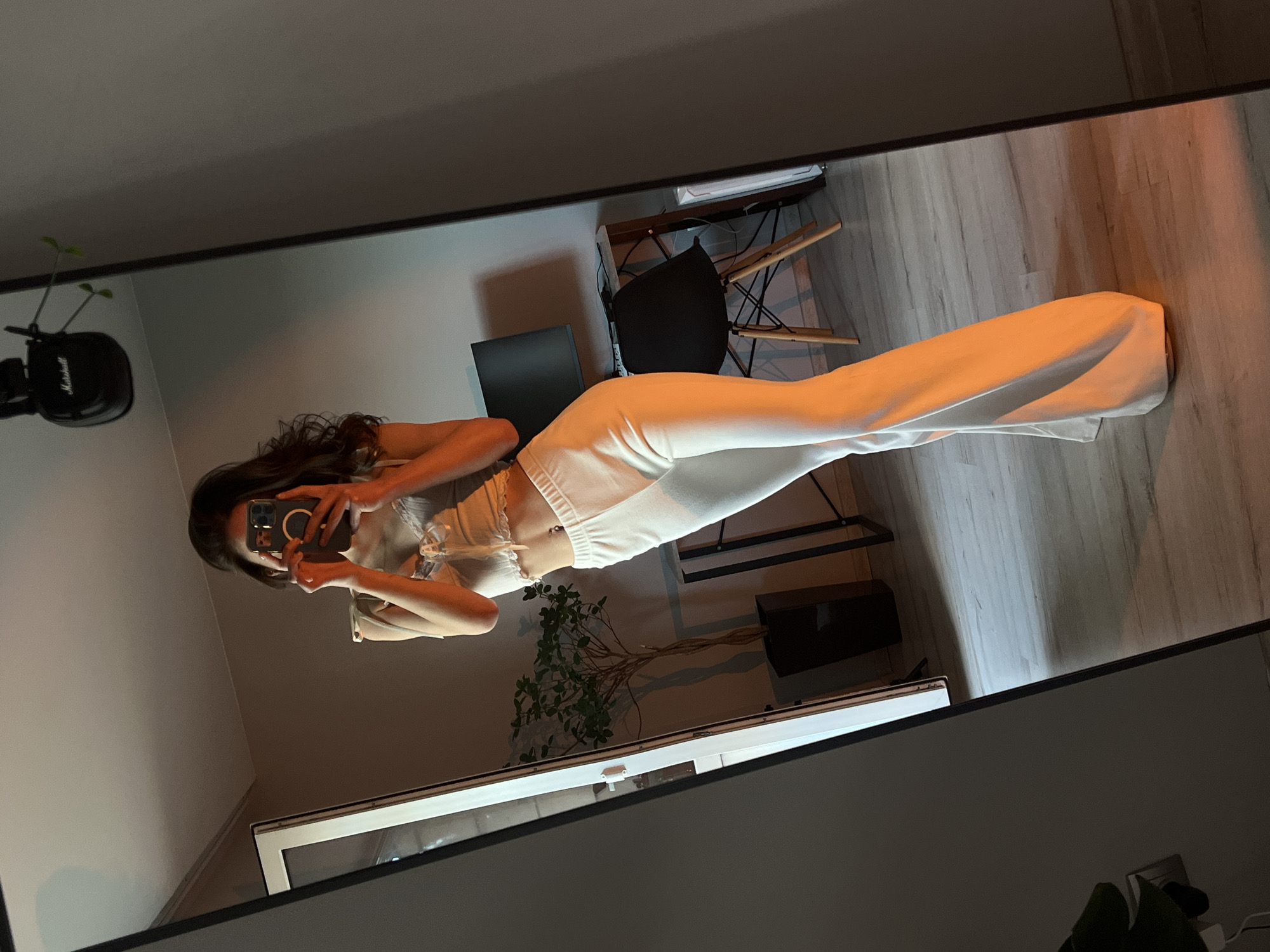 In a room that appears to be in Ternopil, Ukraine, a young woman is taking a mirror selfie. She is wearing a white crop top with lace trim and white flared pants. Her dark hair is long and wavy, and she is holding a smartphone to take the photo. In the reflection of the mirror, a desk with a computer monitor, a black chair, and a tall potted plant can be seen. A pair of headphones hangs from the ceiling in the background. The lighting in the room is warm, with an orange hue casting shadows on the floor.