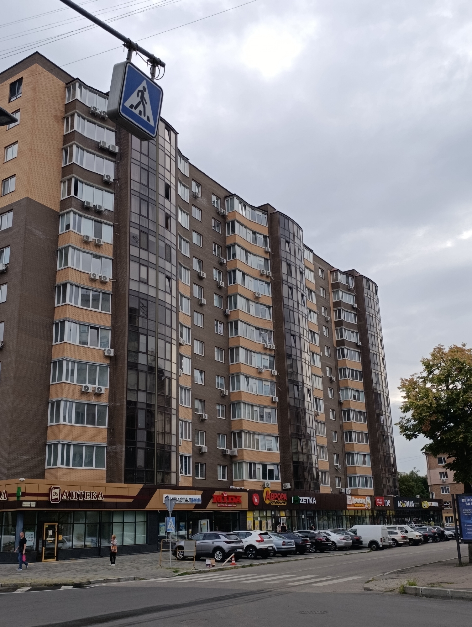 The image shows a multi-story residential building with commercial establishments on the ground floor, located on a street in Cherkasy, Ukraine. The building is characterized by its brick and dark glass facade, with numerous balconies and air conditioning units visible. Several cars are parked along the street in front of the building, and a pedestrian crossing is marked on the asphalt. A pedestrian crossing sign is suspended above the street by a wire. The sky is overcast, suggesting a cloudy day. In the foreground, the asphalt road and sidewalk are visible. In the background, trees and another building can be seen. The commercial establishments on the ground floor have signs in Cyrillic script, indicating businesses such as a pharmacy ("АПТЕКА") and other shops. Two individuals are walking on the sidewalk.