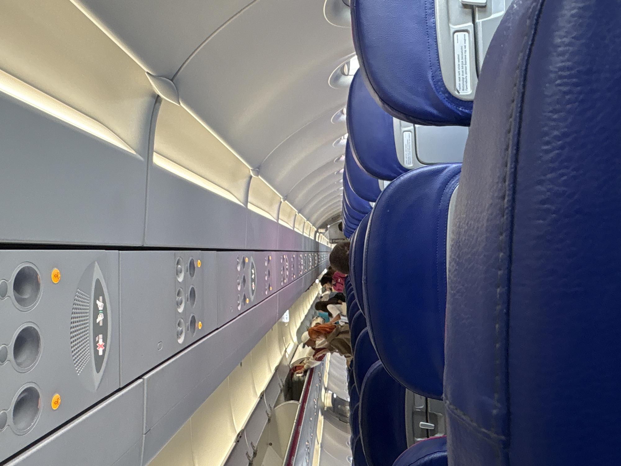 The interior of an airplane cabin is depicted from the perspective of a passenger seated near the aisle. A line of blue leather seats stretches into the distance, partially occupied by passengers. On the left is the cabin wall with overhead compartments, featuring illuminated panels with icons and control buttons. The ceiling is curved with built-in lighting. The image gives no indication of the city of Madrid or Spain.