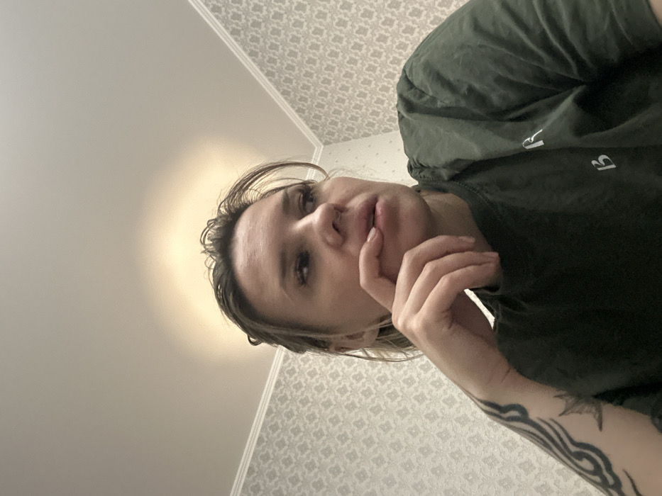 The image presents a low-angle, close-up view of a single adult female in an indoor setting. This environment is consistent with a residential or private interior located in Rostov-on-Don, Russia; however, the precise location within the city cannot be determined from the visual evidence.
The female subject, appearing to be of European descent, occupies the lower half of the frame, gazing upwards and towards the right. Her light-colored hair, possibly damp, is swept back from her face. Notable facial features include visible eyelashes, a small nose ring on her left nostril, and slightly full lips which appear to be unadorned by lipstick. Her right hand is raised, with the index finger and thumb lightly touching her lower lip and chin, suggesting a thoughtful or pensive posture. A distinct dark, patterned tattoo is visible on her left forearm. She is dressed in a dark green t-shirt that displays two white Cyrillic characters, "Я" positioned above "В", on the chest area.
The background consists of a wall covered with light-toned wallpaper featuring a repetitive, geometric or patterned design. A white decorative molding runs along the junction of the wall and a plain, light-colored ceiling. A soft, circular glow of indirect light is visible on the ceiling directly above the subject's head.