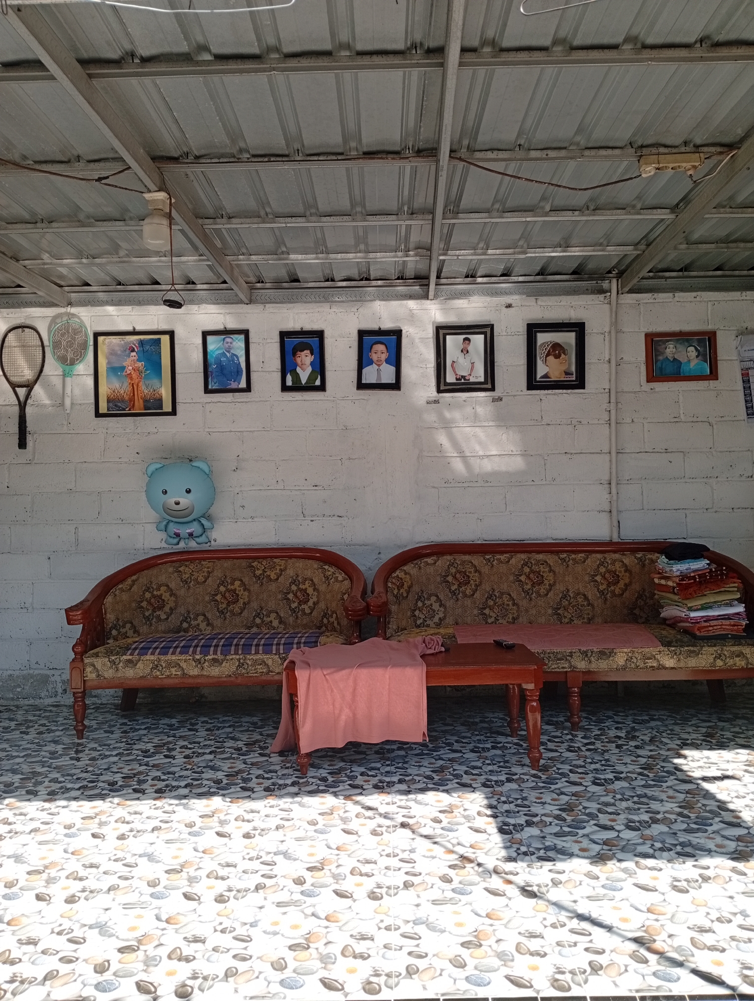 The image depicts an outdoor covered area with a tin roof supported by metal beams. The walls are constructed of white brick. Hung on the wall are several framed portraits, a tennis racket, and a decorative fan. In the foreground, two ornate wooden sofas with patterned upholstery are arranged. A small wooden coffee table sits between them, draped with a pink cloth. To the right of the sofas, a stack of folded fabrics is visible. The floor is tiled with a mosaic pattern resembling pebbles. Shadows from the roof structure cast patterns on the tiled floor, indicating sunlight is present. A blue teddy bear balloon is attached to the wall above the left sofa. The scene appears to be a home's outdoor seating area or patio in Balung, Indonesia.