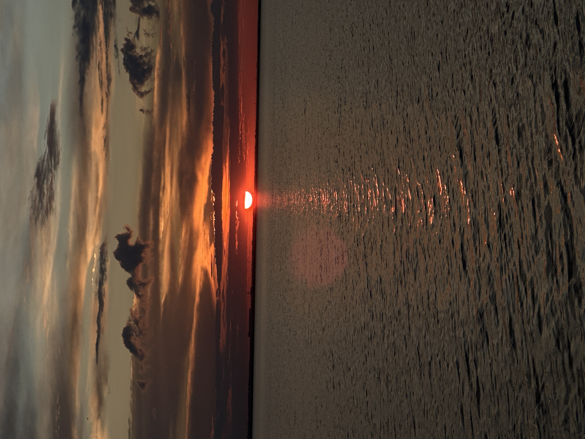A partially obscured sun, low on the horizon, casts a warm orange glow across a body of water. The sky above is a canvas of dramatic clouds, painted with shades of orange, red, and grey, indicating a sunset or sunrise. The water's surface is rippled, reflecting the vibrant colors of the sky. In the distance, a faint, dark line suggests land with some structures, possibly buildings or industrial elements, visible on the horizon. This scene is likely captured on one of the numerous lakes surrounding Kokshetau, Kazakhstan, known for its picturesque natural landscapes, particularly during transitional daylight hours. There are no people or animals visible in the frame.