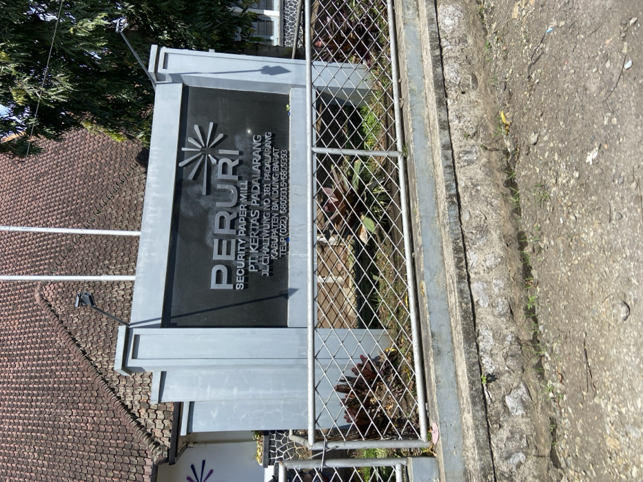 The image shows a sign for "Peruri Security Paper Mill, PT Kertas Padalarang, Jl. Cihaliwung No. 181 Padalarang, Kabupaten Bandung Barat, Telp. (022) 6809315-6809393" at a location with latitude -6.838 and longitude 107.491 in the city of Padalarang, Indonesia. The sign is mounted on a concrete wall with a grey metal fence in front of it. The fence has green vines growing up it. There are trees to the right of the sign. The ground in front of the fence is dirt and gravel. A portion of a building is visible to the left of the sign. The sign has a black background and white lettering. The sign includes a starburst logo above the company name.