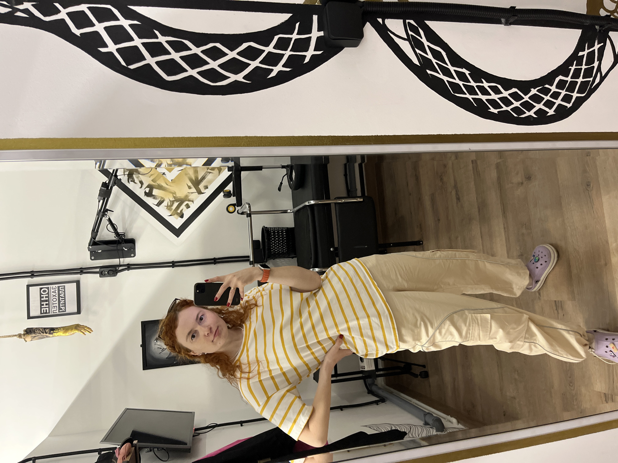 The image shows a selfie taken in a room that appears to be a studio or workspace. The person taking the selfie is a fair-skinned woman with red curly hair, wearing a yellow and white striped shirt and beige cargo pants, and lavender Crocs. She is holding a black smartphone.
In the background of the reflection, there are various objects: a computer monitor, a black office chair, a desk with a camouflage pattern, black electrical conduits running along the walls, framed black and white artwork, and a wall decoration featuring a dangling rubber arm with a yellow glove. Above the mirror is a white wall with a gold accent stripe and abstract black designs.
The location is Chaikovsky, Russia.