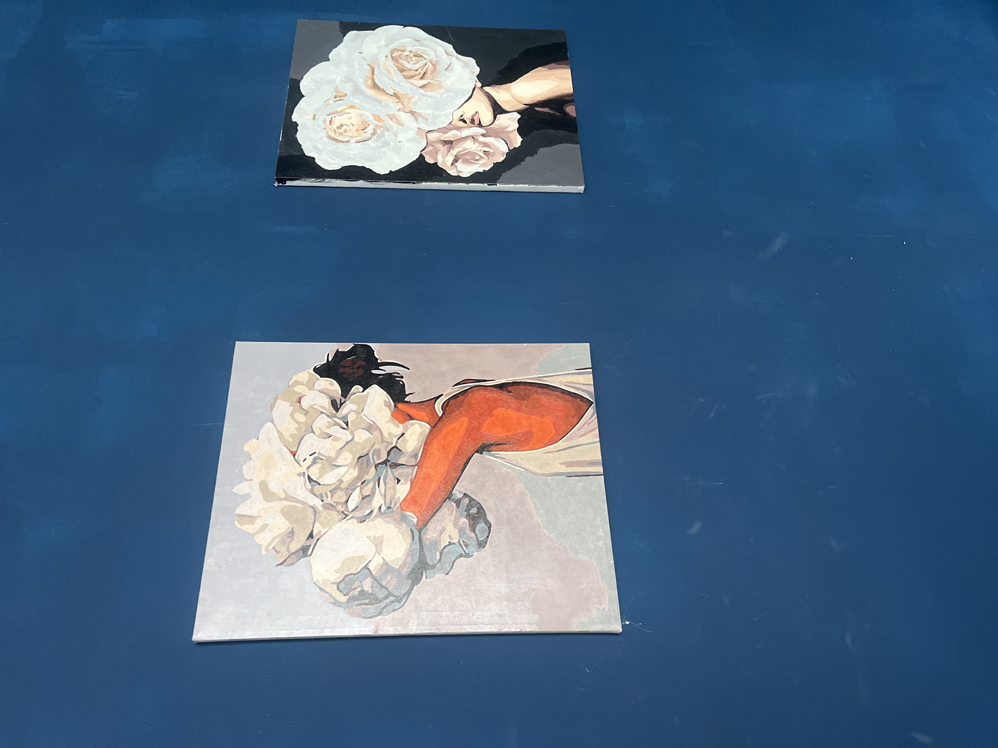 Two square paintings hang on a dark blue wall.
The top painting features a depiction of white and pink roses against a black background, with a beige arm and hand holding the flowers.
The bottom painting portrays a person with dark hair holding white flowers with a copper-colored arm. The background of the second painting is light gray.