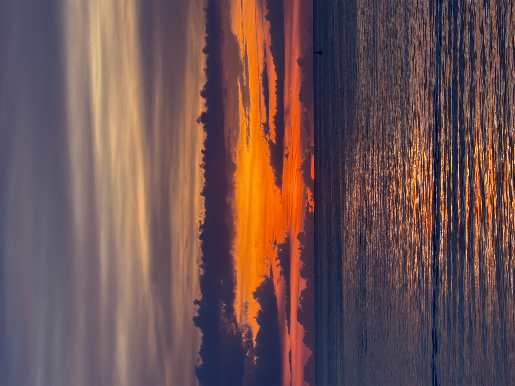 This image depicts a sunset over the ocean, possibly viewed from the coast of ตำบลเกาะพะงัน, Thailand. The sky is ablaze with vibrant colors, ranging from deep purples and oranges to bright yellows and reds, layered by horizontal cloud formations. The sun is setting near the horizon, casting a warm glow across the scene. The ocean is calm, with gentle ripples reflecting the colors of the sky. A few small boats or buoys are visible in the distance on the water. The overall impression is one of tranquility and natural beauty.