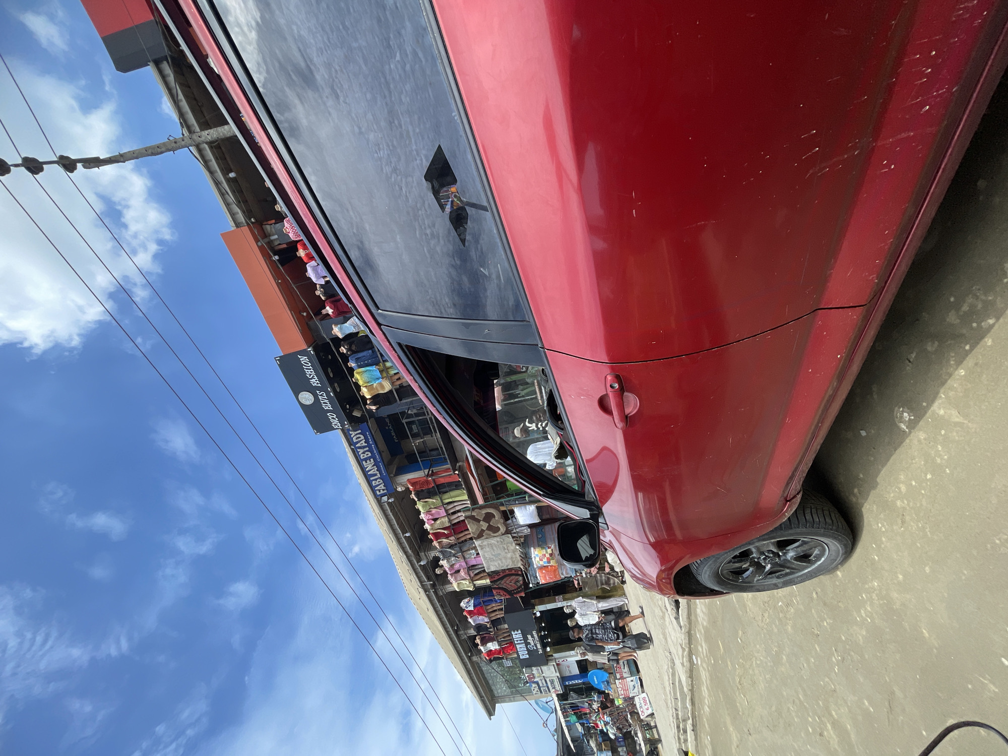 A red sedan is parked on the side of a busy street in a commercial area, likely in Lagos, Nigeria, under a bright blue sky with scattered clouds. The most notable detail is the significant damage to the car's rear passenger window, which is shattered with a distinct jagged hole, suggesting a break-in or vandalism. The red paintwork of the vehicle also displays multiple scratches and scuff marks, particularly on the rear passenger side.
In the background, a lively street scene unfolds with several people visible, some walking and others standing near shops. The shops display various goods, including clothing and rugs, indicating a market or retail district. Overhead, multiple power lines are visible, connecting to utility poles.
Visible text from the shop signs and banners includes "FABLANE BY ADY," identified as a "Unisex Clothing Store," and "ENCO ELVIS FASHION." Another sign reads "BURN FIRE" along with a phone number, "Tel 00037216895." A "DSTV" logo is also visible, and a sign near a vendor shows "6.500" and "POS," possibly indicating a price or point-of-sale service. The scene depicts a typical, bustling urban environment, with the damaged car being the focal point.