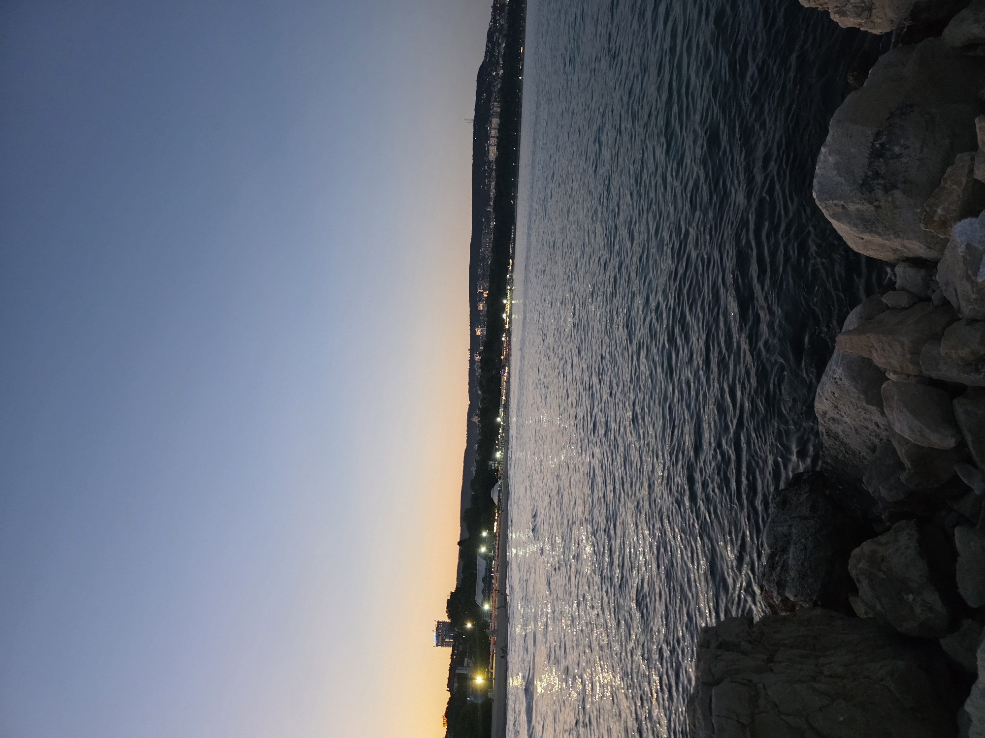 The image depicts a coastal scene at dusk in Varna, Bulgaria. In the foreground, a rocky shore leads to the dark, rippling waters of the sea. The water reflects the fading light of the sky and the distant city lights. Along the coastline in the midground, a strip of beach is visible, dotted with beach umbrellas and illuminated by numerous lights from buildings and structures. Beyond the beach, the Varna cityscape stretches across a hill, with buildings and lights creating a distinct urban silhouette against the twilight sky. The sky itself transitions from a pale orange hue near the horizon, indicative of sunset, to a deep blue overhead. The overall impression is one of a serene evening by the sea in a bustling coastal city.