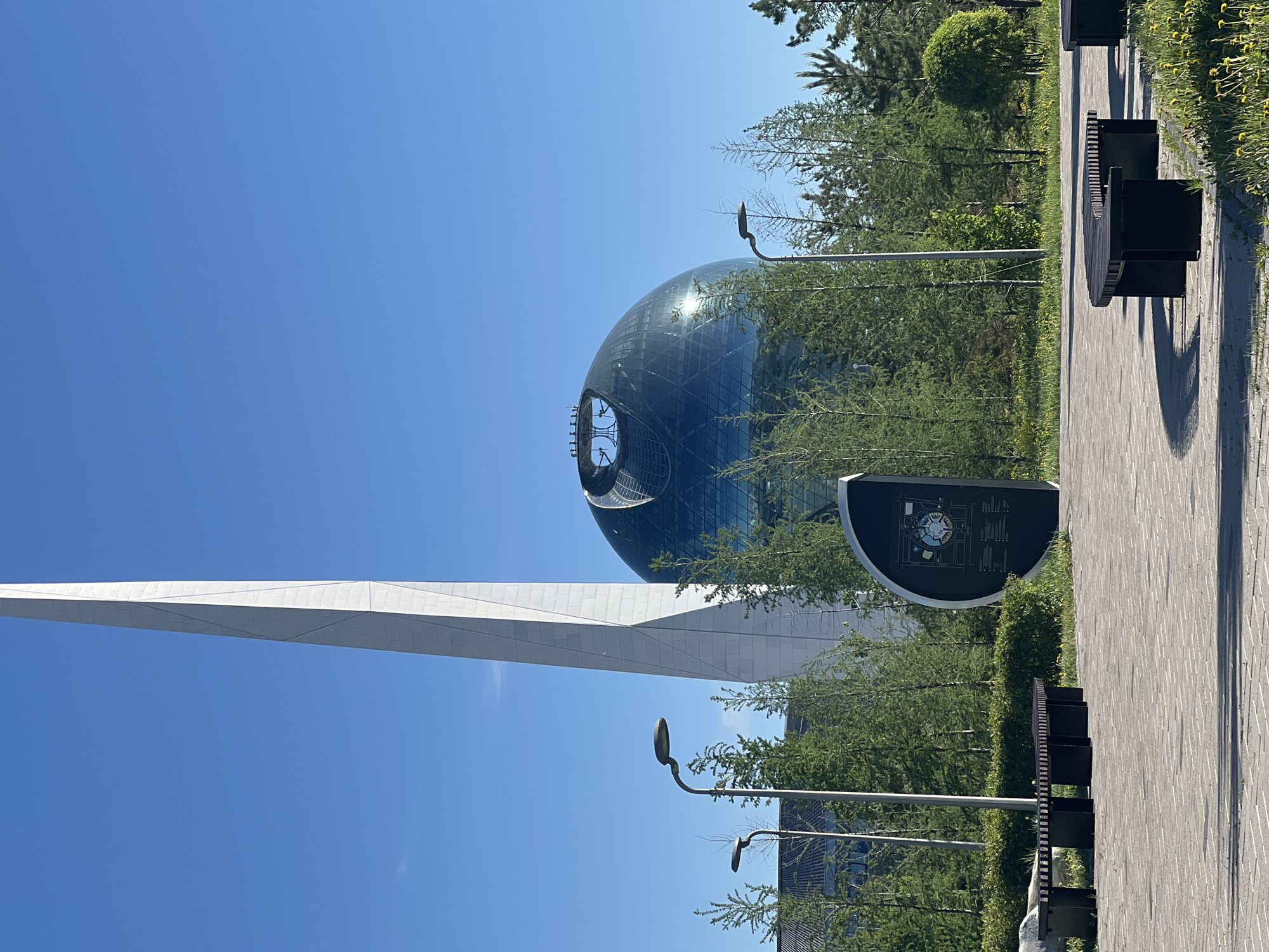 The image shows a view in Astana, Kazakhstan, featuring the Nur Alem sphere and a white, obelisk-like structure against a clear blue sky. The Nur Alem sphere is a large, glass-covered structure with a noticeable opening at the top. The obelisk is white and triangular, pointing upwards. A paved pathway with dark-colored benches is visible on the right side of the frame, along with greenery consisting of trees and bushes. A black information board stands near the pathway. Street lamps are positioned among the trees.
