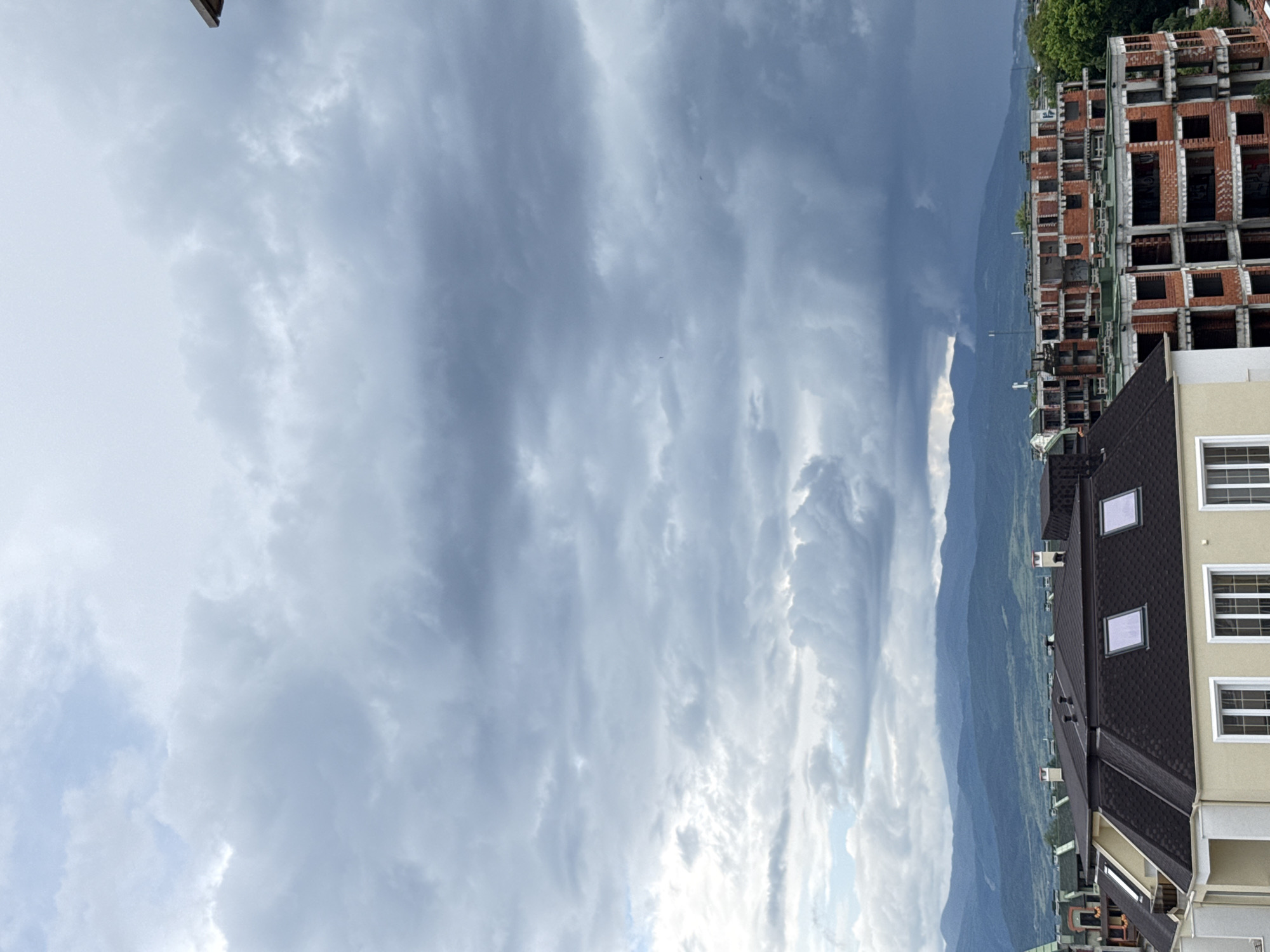 The image shows a landscape with a cloudy sky occupying most of the frame. A mountain range is visible in the middle ground, with a valley or town nestled at its base. A building with multiple windows and a dark roof is on the right side of the frame. Further to the right, a partially constructed brick building can be seen. The image does not contain enough detail to determine whether the location is in Plovdiv, Bulgaria. There are no visible people, subjects, actions, or interactions.