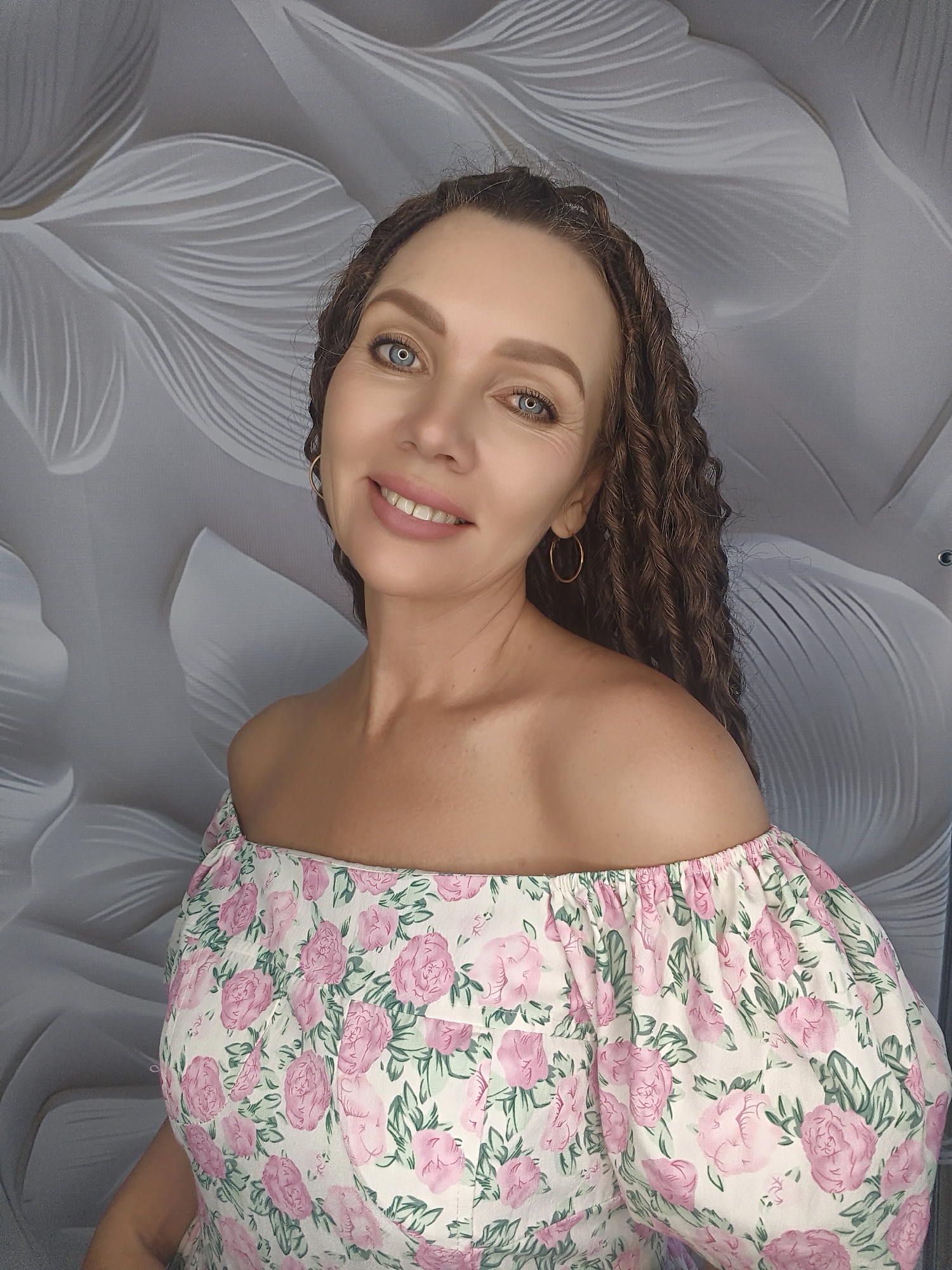 A medium shot captures a woman with long, curly brown hair, light blue eyes, and a warm smile against a backdrop of a grey, abstract floral pattern. She is wearing an off-the-shoulder white dress adorned with pink roses and green leaves. The woman is positioned centrally in the frame, looking directly at the camera. She has on simple gold hoop earrings.