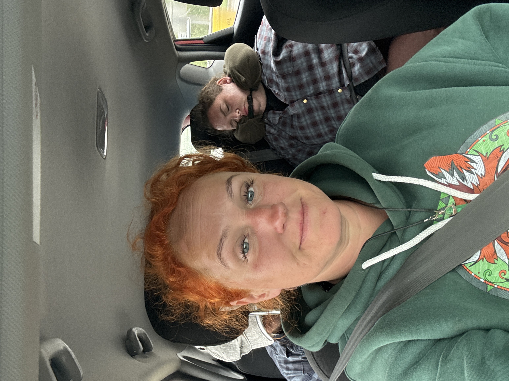 The image depicts the interior of a vehicle, likely a car, where two individuals are present. The person in the foreground, with bright red, curly hair, is looking directly at the camera and appears to be the driver. They are wearing a green hooded sweatshirt with a white drawstring and a dark necklace. Their left shoulder is angled towards the camera. To the right and slightly behind, another individual is visible, sleeping. They are wearing a plaid shirt and have a travel neck pillow around their head, with a strap across their chin. A third person, wearing a hat and a plaid shirt, is partially visible in the background, seated behind the driver. The interior of the car shows the ceiling, a seatbelt, and the back of a headrest. The lighting suggests it is daytime, and the scene appears to be set within or near the city of Tatarstan, Russia.