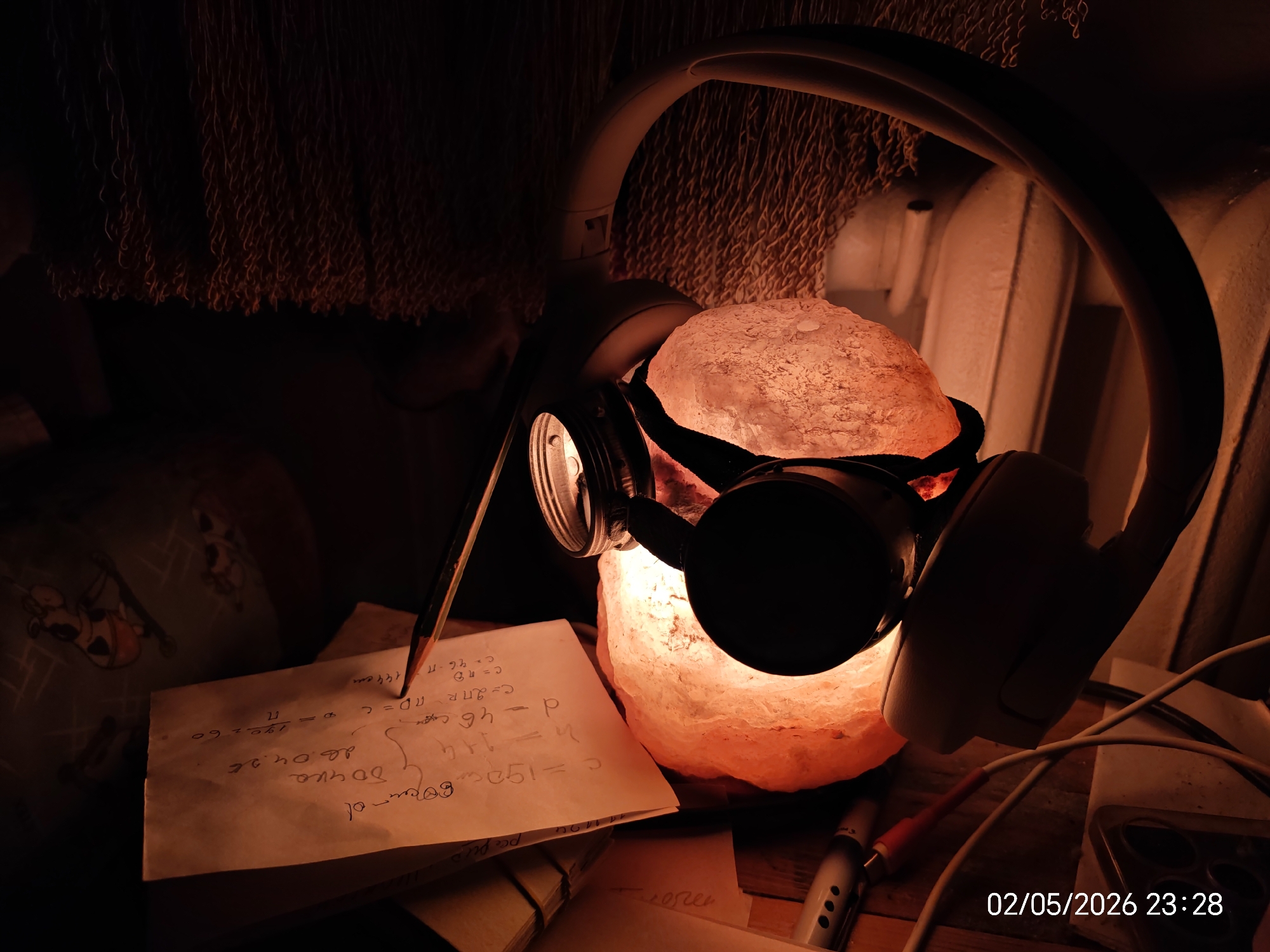 The image shows a desk cluttered with various items, bathed in the warm, orange glow of a Himalayan salt lamp. A pair of white headphones rests on and around the lamp, with the right earcup prominently visible. To the left of the lamp is a piece of paper with handwritten notes, likely mathematical formulas or equations, along with a pencil resting on top. Cables, a pen, and other unidentified objects are scattered across the desk surface. The date "02/05/2026 23:28" is visible in the bottom right corner of the image. The location is unknown, as no identifying information is present.