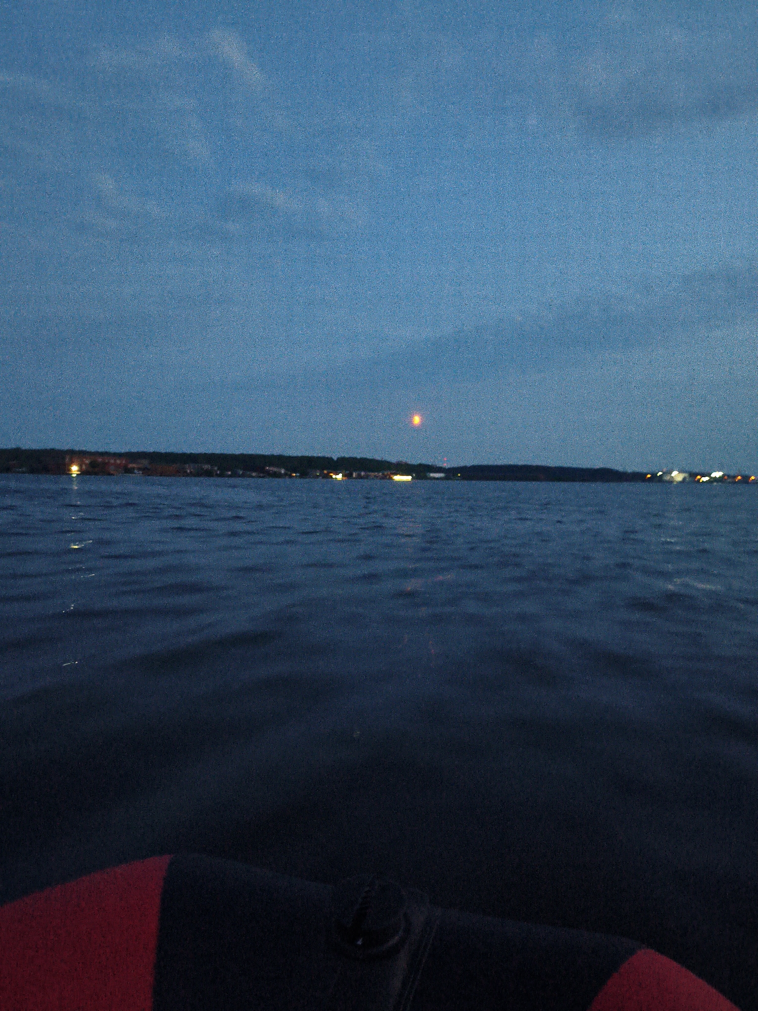 The image captures a serene twilight scene over the Kama River in Chaikovsky, Russia. The sky is a deep, dusky blue, with faint clouds visible. A distant, hazy orange light, possibly the moon or a reflection, hangs in the sky. The river is calm, with gentle ripples reflecting the dim light. Along the far bank, a line of trees and distant city lights form a dark silhouette against the horizon. In the foreground, the edge of an inflatable boat, with its red and black material, is visible, suggesting the photo was taken from the water. The overall atmosphere is peaceful and tranquil.