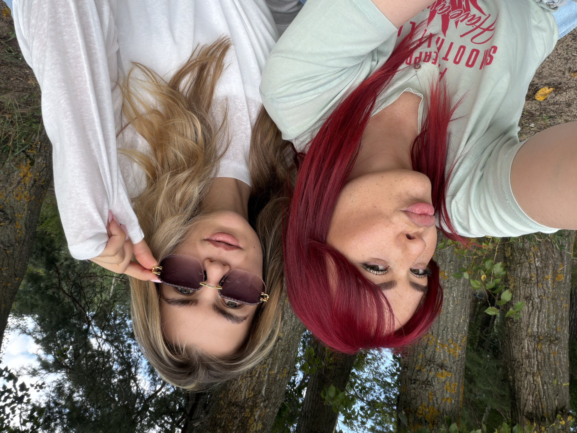 Two women are taking a selfie outdoors, with trees and foliage in the background. The woman on the left has bright red hair, freckles, and is wearing a light green t-shirt with red text. She is puckering her lips for the photo. The woman on the right has blonde hair and is wearing sunglasses and a white top. She is adjusting her sunglasses. The location is identified as the village of Uzyukovo, Russia, which is characterized by its natural, wooded environment.