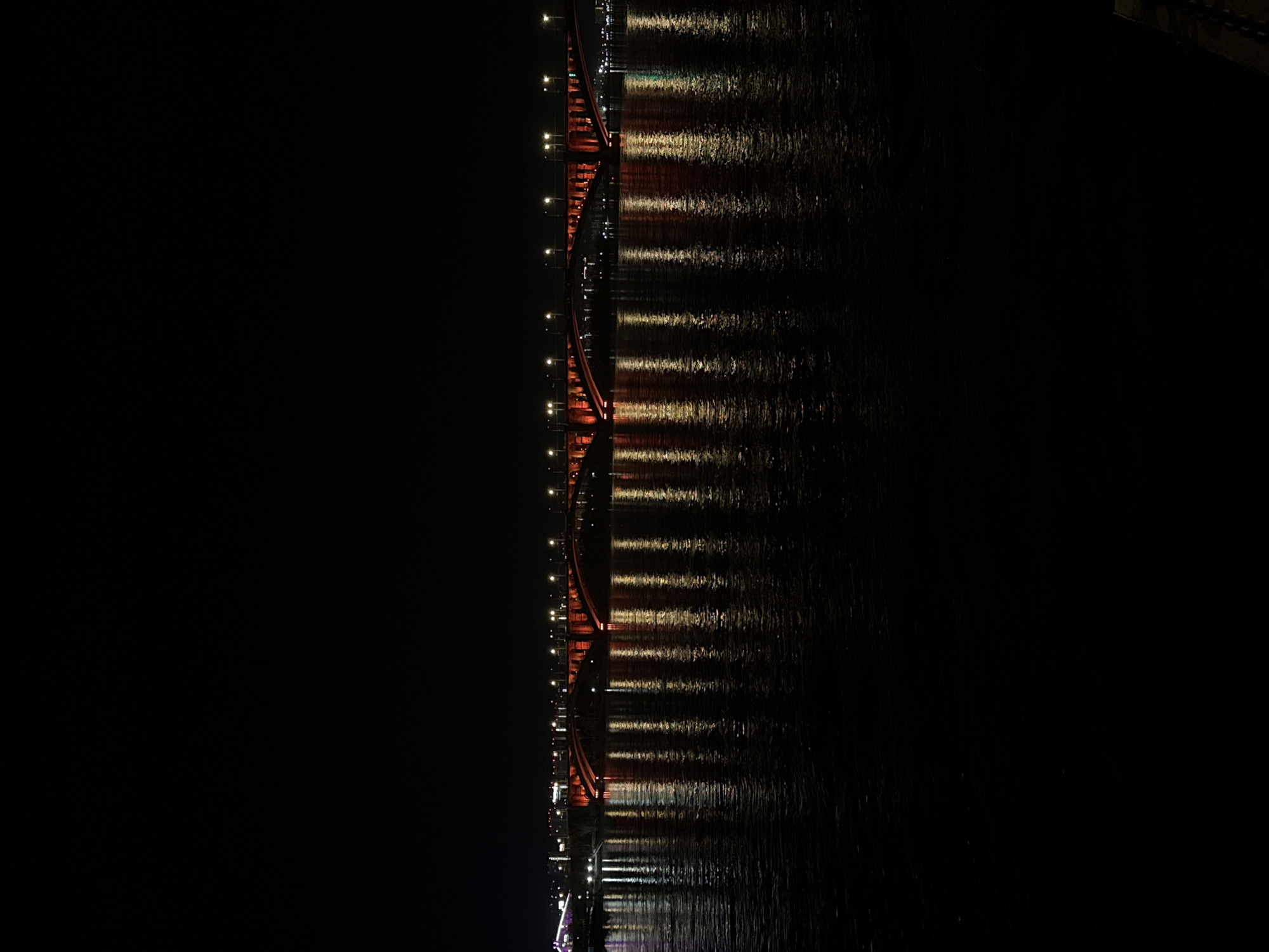 This media file presents a nighttime view of a prominent, red-colored bridge stretching across a body of water, captured in what appears to be a rotated orientation (originally landscape, rotated 90 degrees counter-clockwise). The bridge occupies the central vertical axis of the image, showcasing its intricate metal truss or arch structure illuminated by numerous bright lights. These lights, possibly streetlights or architectural lighting, cast striking, elongated reflections onto the dark, rippling water surface to the right of the bridge. The reflections appear as shimmering streaks of golden-yellow and reddish-orange light, mirroring the bridge's vibrant color and the individual light sources.
The left side of the image is dominated by the absolute darkness of the night sky, providing a stark contrast to the illuminated bridge. The water on the right exhibits subtle textures from gentle ripples, enhancing the visual effect of the light reflections. There are no visible people, vehicles, or specific activities taking place on the bridge or water, conveying a sense of stillness and tranquility. The clear and distinct reflections suggest calm weather conditions without heavy rain or fog. Given the context of Krasnoyarsk, Russia, the distinctive red hue and structural design strongly suggest this is the famous Krasnoyarsk Railway Bridge (also known as the Yenisei Bridge). No visible text can be discerned in the image.