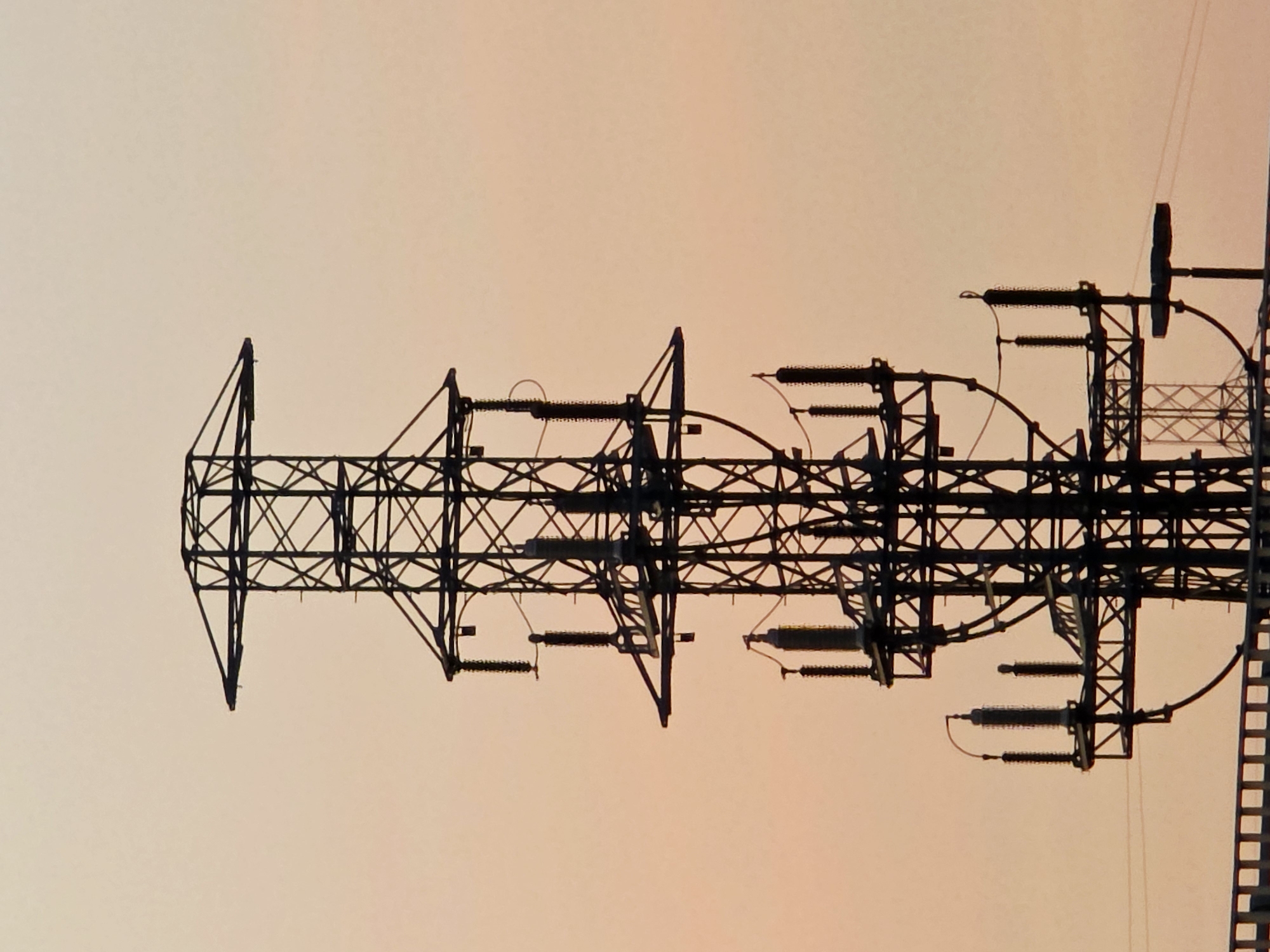 This is a close-up, silhouette photograph of the top section of a large, industrial electricity pylon against a soft, gradient sky that transitions from a pale yellow at the bottom to a light pink at the top. The pylon is a complex structure made of dark metal lattice work, featuring multiple horizontal crossarms from which insulators and electrical conductors are suspended. Several large, dark, cylindrical insulators are visible, connected by wires. The pylon's design is tiered, with wider sections at the top and middle, tapering slightly towards the bottom of the frame. The overall impression is one of industrial infrastructure against a muted, atmospheric sky. The image does not depict any people, animals, or specific landmarks that would indicate a location in Warsaw, Poland.