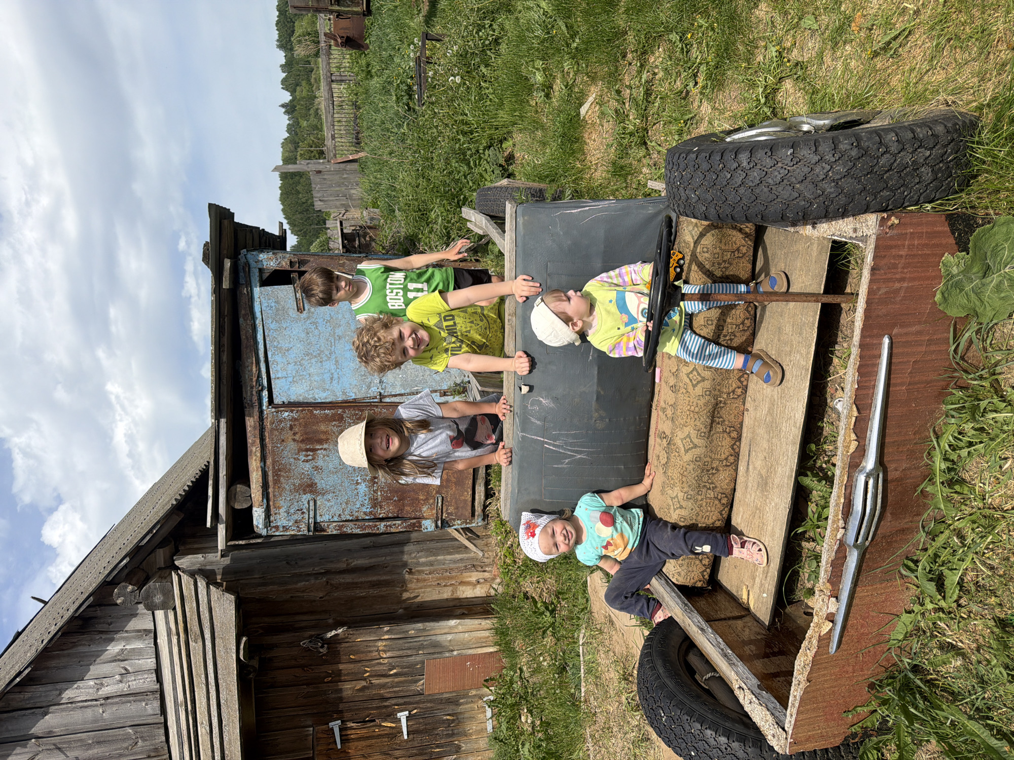 Five children are playing on a makeshift play vehicle outdoors. The vehicle appears to be constructed from various materials, including tires, wood, a black cylindrical container, and furniture cushions. The children range in age and are wearing casual clothes. They are positioned in and around the vehicle. Behind the vehicle is an old, weathered wooden and corrugated metal structure, possibly a shed or outbuilding. The setting appears to be in a rural or semi-rural area, with grass and foliage visible. Based on the prompt information, the location is Panteleevka, Russia.