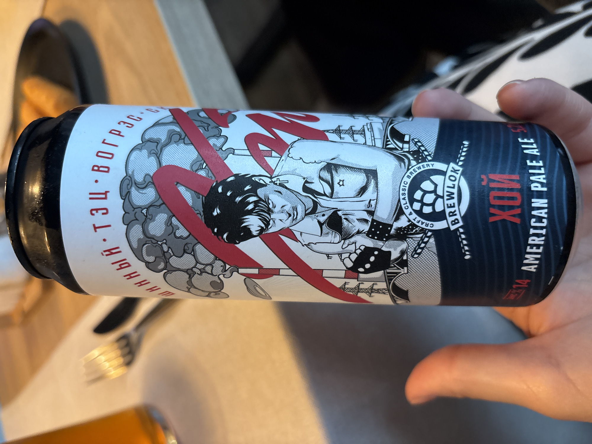 The media file shows a close-up of a person's hand holding a can of beer. The can has a black background with a colorful, graphic design featuring a stylized image of a person with dark hair, a red ribbon, and various industrial or abstract elements. Text on the can includes "ИННЫЙ ТЭЦ ВОГРЭС-", "BREWLOKO", "ХОЙ", and "AMERICAN PALE ALE". The scene appears to be indoors, possibly at a table, as a fork and knife are visible in the blurred background, along with a wooden surface. The lighting suggests it could be daytime, but there are no explicit indicators of time of day or weather. The context "Novaya Usman', Russia" suggests the location where the image was taken. The focus is clearly on the beer can, with the hand serving to frame and present the object.