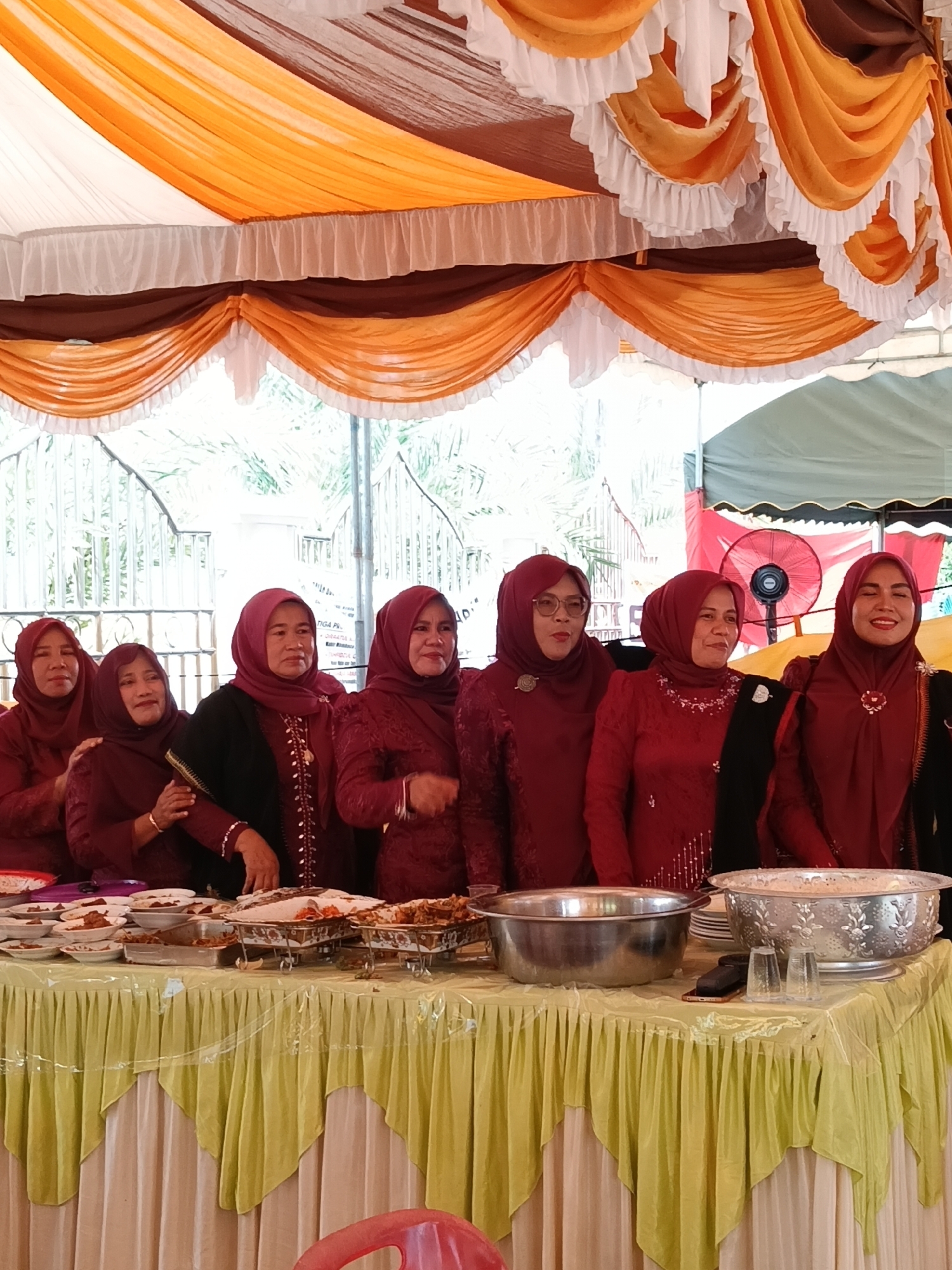 The image captures a group of seven women standing behind a table laden with food. The setting appears to be an outdoor event in Lhokseumawe, Indonesia, as evidenced by the tropical greenery visible in the background. The women are uniformly dressed in maroon attire, including headscarves, which suggests a coordinated effort or participation in a specific function. The table is covered with a yellow and white skirted tablecloth, and a variety of serving dishes and bowls are placed on top. The table holds platters of food, as well as two large metal bowls. Overhead, a decorative canopy with yellow, brown, and white fabric drapes create a festive atmosphere. A red electric fan is slightly visible in the background to the right of the women.