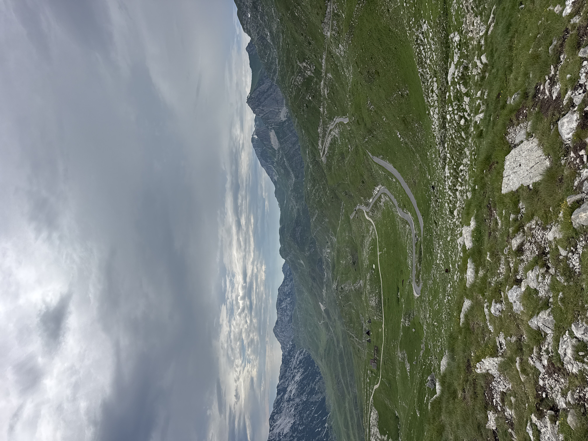 The image shows a panoramic view of a steep, grassy mountainside with a winding road carved into it. The road is paved and appears to have multiple switchbacks as it ascends the mountain. Several small, dark-roofed buildings are visible along the road, suggesting a small settlement or collection of chalets. The terrain is rugged, with large rocks interspersed with the vibrant green grass. In the distance, a sheer, rocky mountain face rises, its slopes showing patches of snow. The sky above is filled with dramatic, white and grey clouds, with areas of bright sunlight breaking through, indicating a dynamic weather pattern. The scene suggests an outdoor recreational area, possibly a hiking or scenic driving destination. The time of day appears to be daytime, with the lighting suggesting either mid-morning or late afternoon. The overall mood is one of natural beauty and ruggedness. There is no visible text in the image.