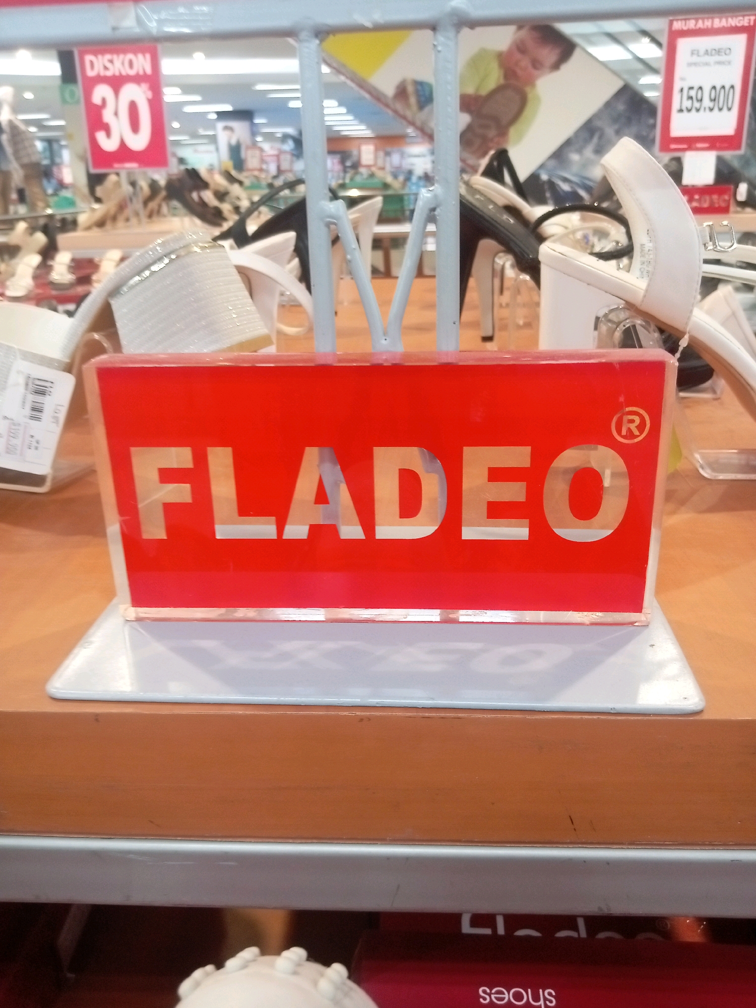The image depicts a retail store environment, likely a shoe store, in Batam, Indonesia. In the foreground, a red sign with the brand name "FLADEO" is prominently displayed. The sign has cut-out letters showing the background. To the left of the sign, a price tag with "DISCOUNT 30%" is visible, indicating a sale. Behind the sign, several pairs of women's shoes, including white high-heeled sandals and black heels, are arranged on display shelves. In the background, a large promotional image of a child holding a shoe can be seen. The overall scene suggests a commercial setting focused on footwear sales.