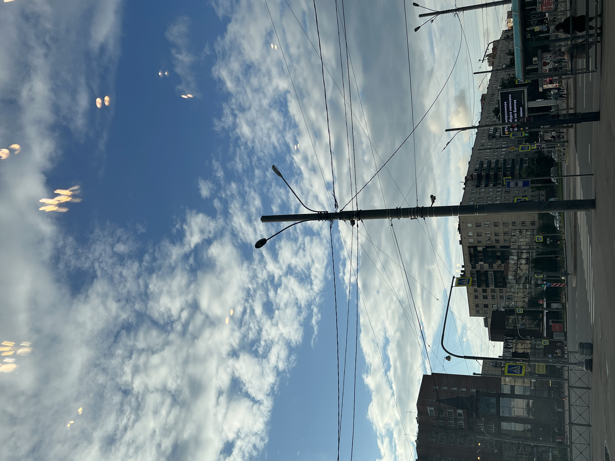 This is a street-level, eye-level, medium shot photograph taken in Sinyavino, Russia, depicting a cloudy sky with blue patches over a city intersection. Several streetlights and a cluster of overhead wires dominate the mid-ground. In the background, multi-story apartment buildings line the streets, with the left side of the frame featuring a darker brick building and the right side showing lighter-colored apartment blocks. Pedestrians are visible walking on sidewalks and waiting at a bus stop. Traffic lights are present, and a crosswalk is marked on the road. The overall impression is an urban street scene under a dynamic sky, suggesting it might be late afternoon or early evening due to the lighting.
