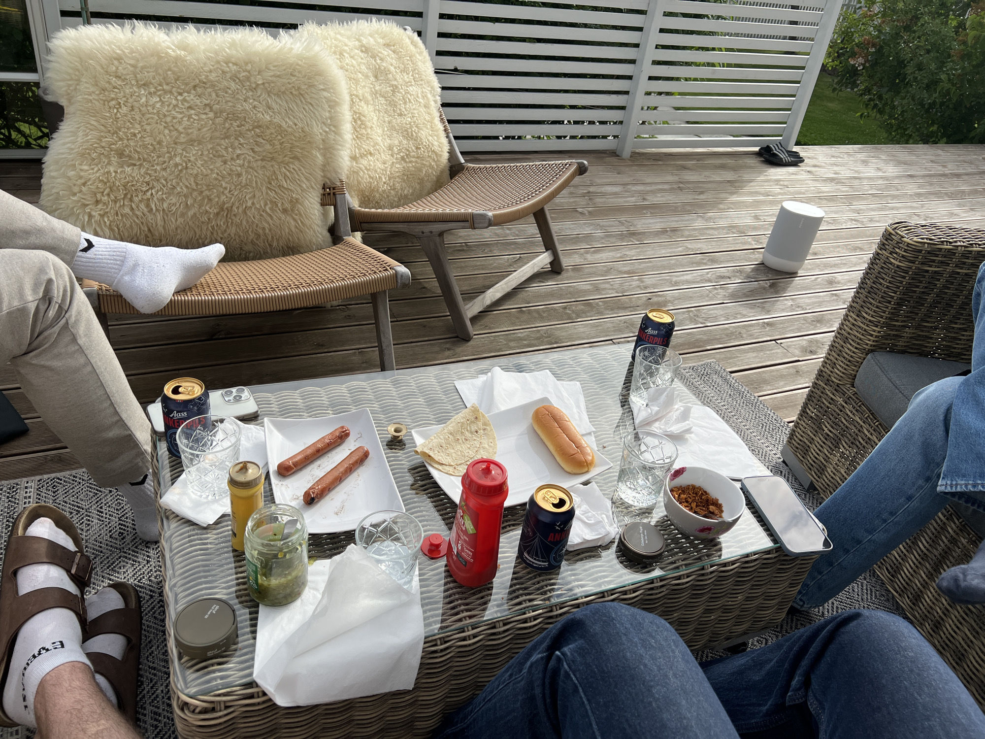 The image depicts a casual outdoor gathering on a wooden deck, likely during daytime and pleasant weather, judging by the bright sunlight casting shadows. The setting appears to be a patio or balcony, with a white slatted fence in the background and greenery visible beyond. The overall mood is relaxed and convivial, suggestive of a social event like a barbecue or a casual meal.
Several people are present, though their faces are not clearly visible. The focus is on their legs and feet, dressed in casual attire like jeans, track pants, and socks. One person is wearing brown Birkenstocks with white socks, another is in white socks and track pants, and two others are in blue jeans. Two plush, cream-colored sheepskin cushions rest on wicker chairs, adding a touch of comfort and style.
On a glass-topped wicker coffee table, a spread of food and drinks is laid out. There are two cooked sausages on a white plate, a hot dog bun, and a piece of flatbread. Condiments like ketchup and mustard are visible, along with a jar of pickles. Several glasses, some filled with water, and cans of "Aass Pils" beer are also on the table. A white smartphone rests on the table, alongside a small bowl containing what appears to be fried onions. A cylindrical white speaker is placed on the wooden deck further back. The presence of food, drinks, and comfortable seating suggests an informal meal or gathering. The location context of Larkollen, Norway, places the scene in a coastal area known for its summer recreational activities.