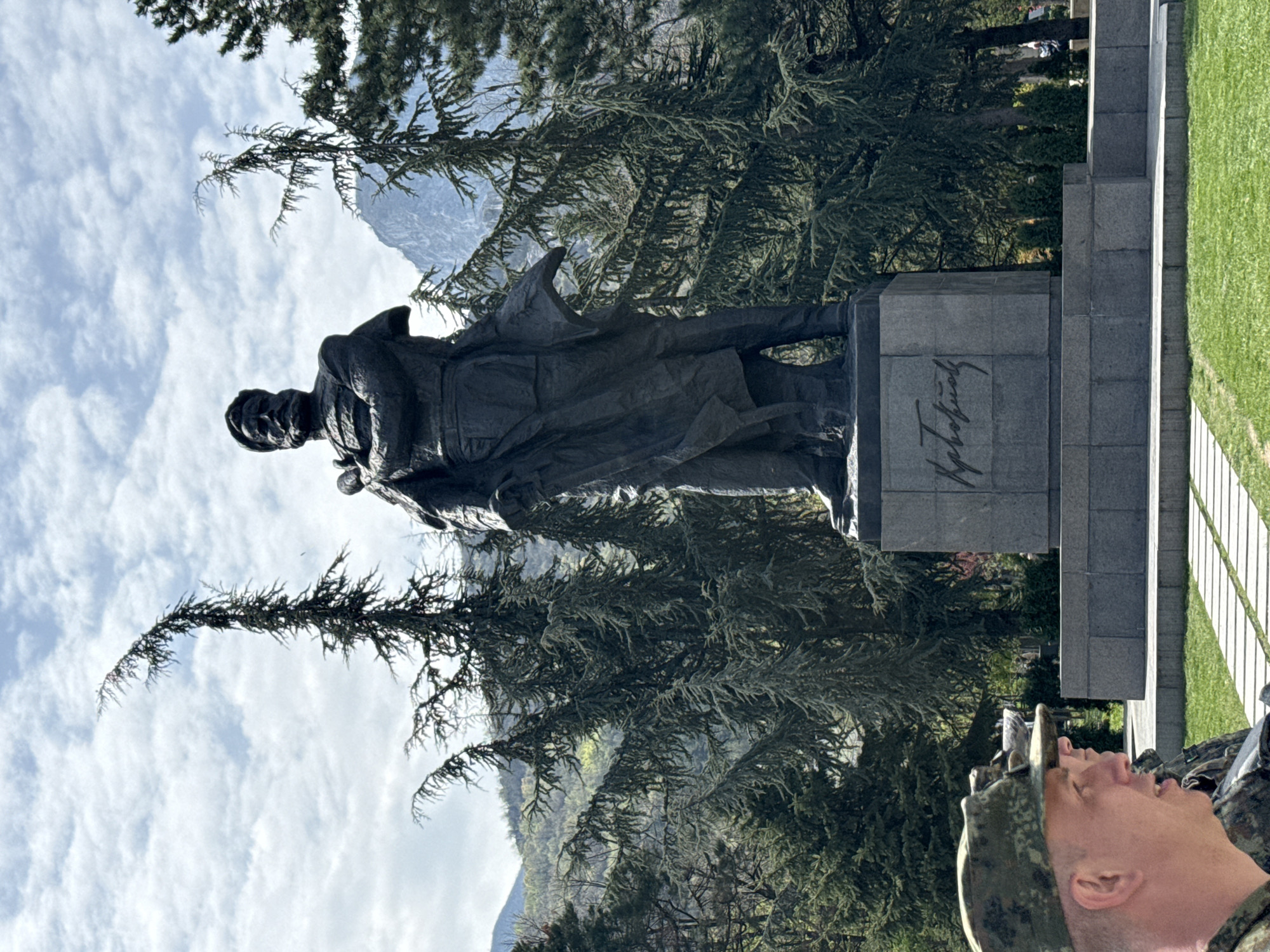 A bronze statue stands on a stone plinth amidst greenery. The statue depicts a man in a long coat and hat. Cyrillic script is inscribed on the plinth. Trees frame the statue. A partly cloudy sky is visible. A person is visible in the bottom right corner. Based on the Cyrillic script and other details, the location is likely in Vratsa, Bulgaria.
