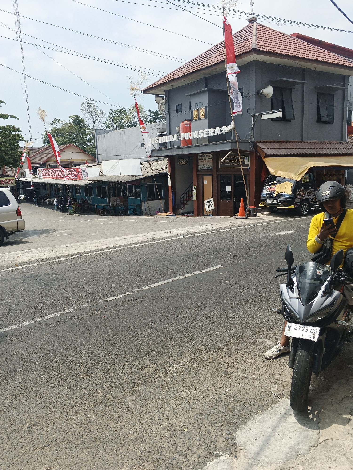 The image shows a street scene in Pacet, Indonesia. In the foreground, a person wearing a yellow shirt and a helmet is sitting on a motorcycle and looking at their phone. The motorcycle has a license plate that reads "W 2793 CQ". To the left of the motorcycle is a road with a white line. In the background, there are several buildings, including a gray building with a red tiled roof and flags displayed. One of the flags is red and white, resembling the Indonesian flag. There are also some stalls or small shops with blue awnings. A silver car is parked on the left side of the road. Trees and utility poles are visible in the background. The sky is overcast.