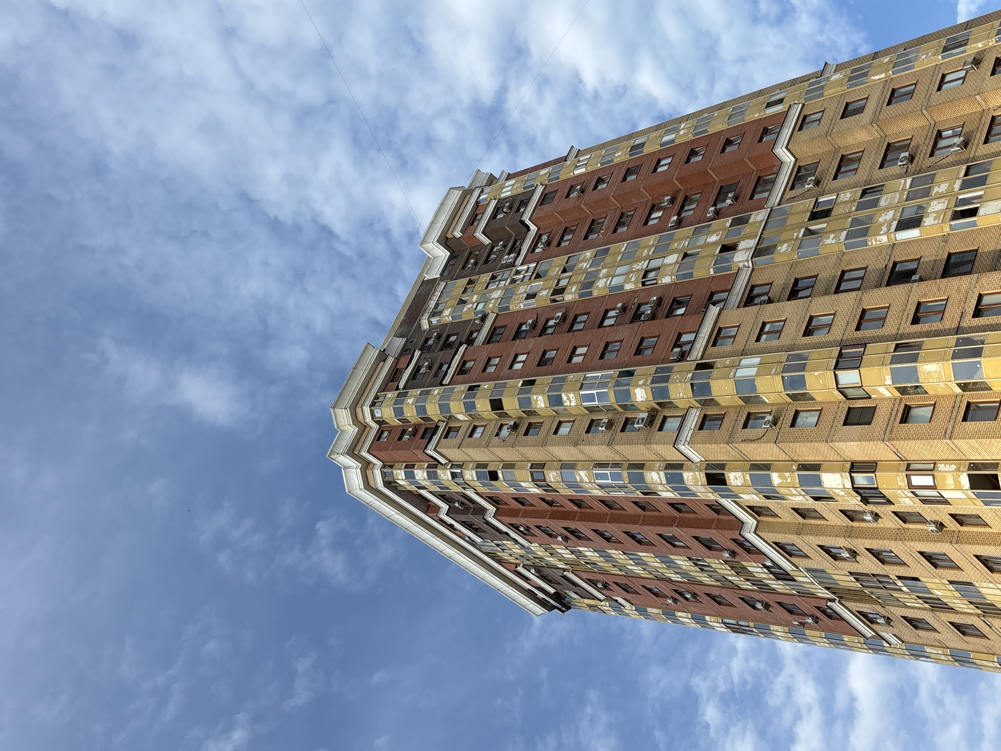An extreme low-angle outdoor medium shot captures a tall, multi-story residential building under a clear blue sky with scattered white clouds. The building, a prominent feature of the Moskovskiy, Russia cityscape, is constructed with alternating bands of red brick and yellow-gold paneling. Numerous windows, many of which are tinted or reflective, are visible across the facade. Air conditioning units are mounted beneath many of the windows. The building has a stepped roofline with white decorative trim. Two thin wires, likely power or communication lines, stretch across the upper portion of the sky.