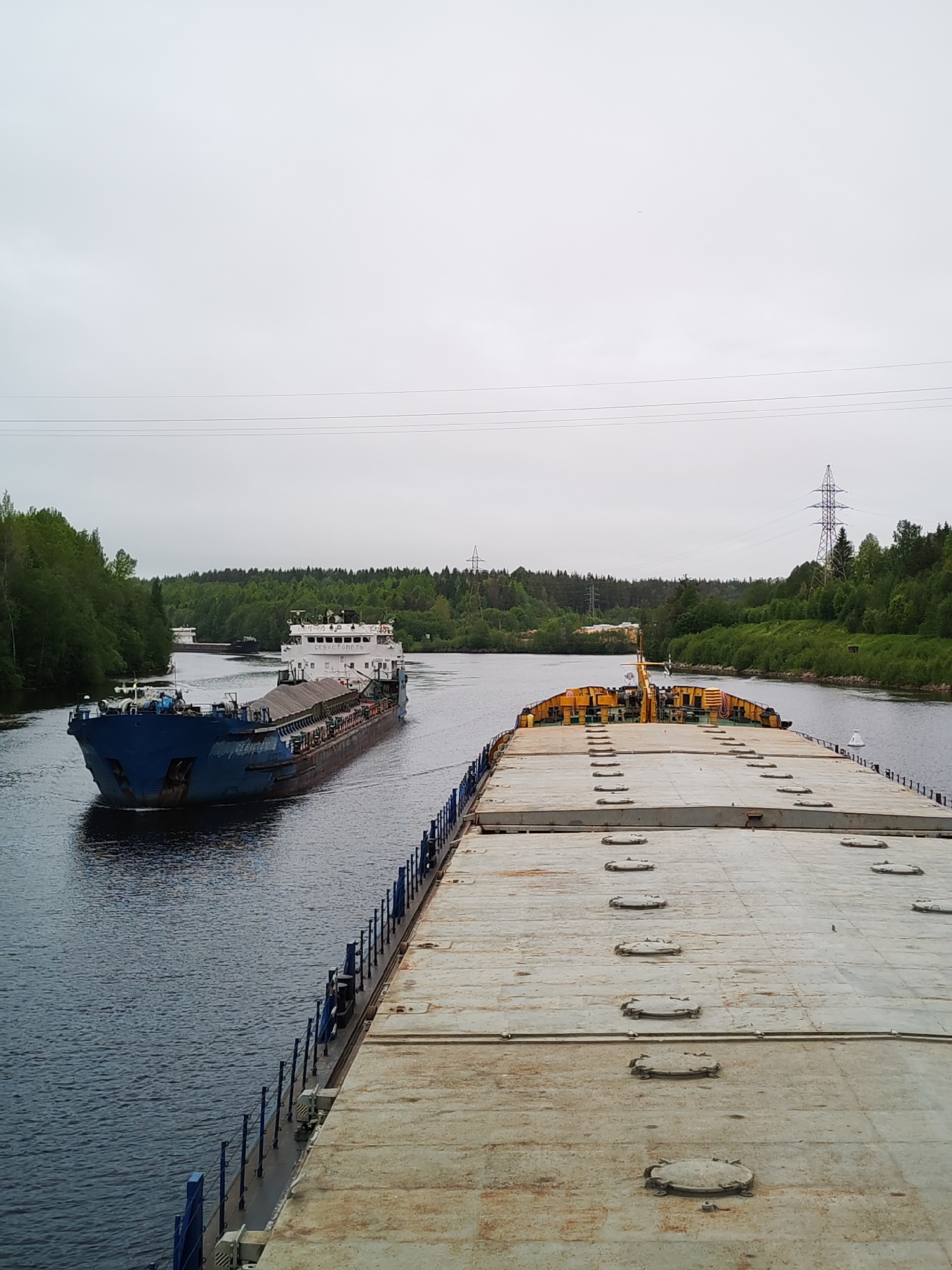 The image depicts a waterway in Depo, Russia, where two large barges are navigating the channel. The foreground barge is mostly gray with several circular hatches. The second barge is dark blue. Trees and power lines line the banks of the channel. The sky is overcast.