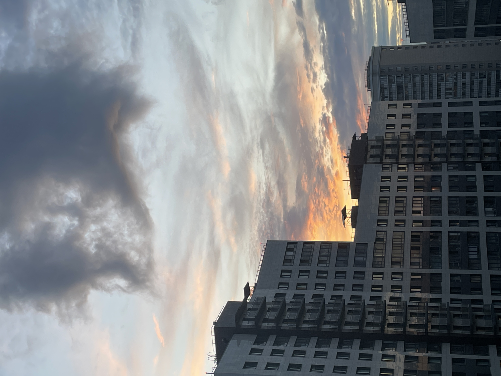 This is a vertically oriented photograph of a cityscape at dusk. The primary subjects are modern, multi-story buildings with numerous windows and balconies. The architecture suggests an urban setting, possibly residential or office complexes. The sky dominates the upper left portion of the image and displays a dramatic sunset with a mix of dark, heavy clouds and lighter, wispy clouds illuminated by the setting sun, casting hues of orange, pink, and yellow. The buildings are silhouetted against this colorful sky, with some illuminated by the fading light. The overall scene evokes a sense of urban grandeur and the fleeting beauty of a sunset. There is no visible text in the image.