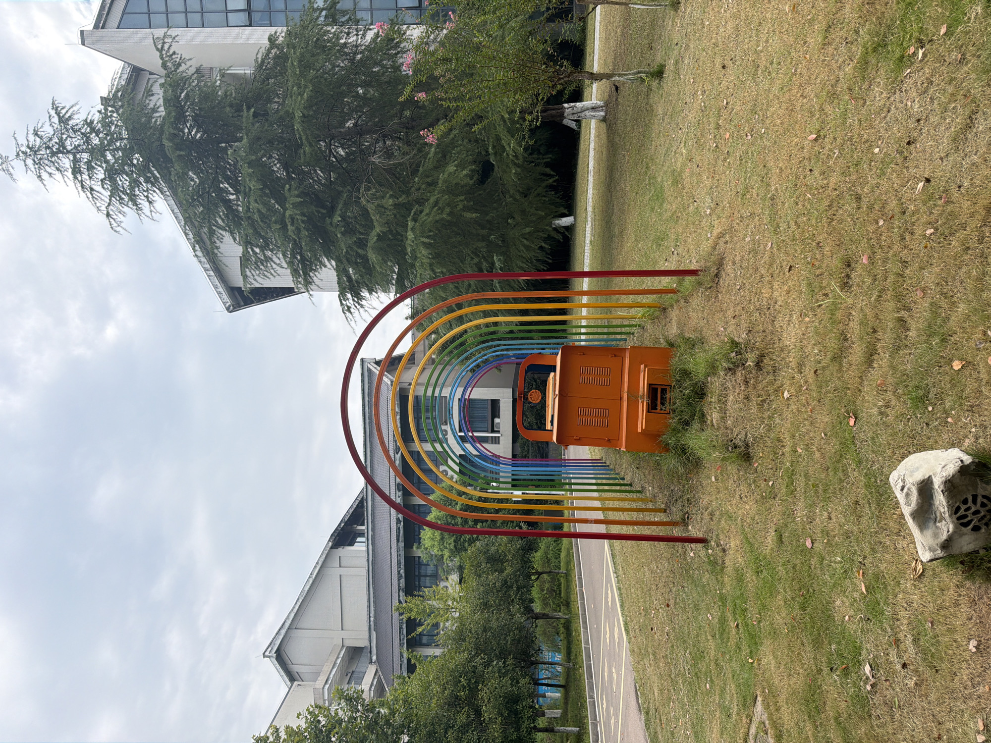 This is an outdoor, eye-level, full shot of a colorful rainbow archway structure in Jiangjin, China. The archway, made of metal poles painted in a rainbow of colors, frames an orange, miniature train-like structure. The train is positioned in the center of the grassy area, between the archway and a paved path on the left. The background features a large evergreen tree on the right, alongside a modern building with glass windows and brick accents. To the left of the evergreen tree and building, another building with a dark roof and lighter walls is visible. The ground is covered in grass, with some fallen leaves scattered around, and a gray, decorative rock is in the foreground. The sky is overcast with visible clouds.