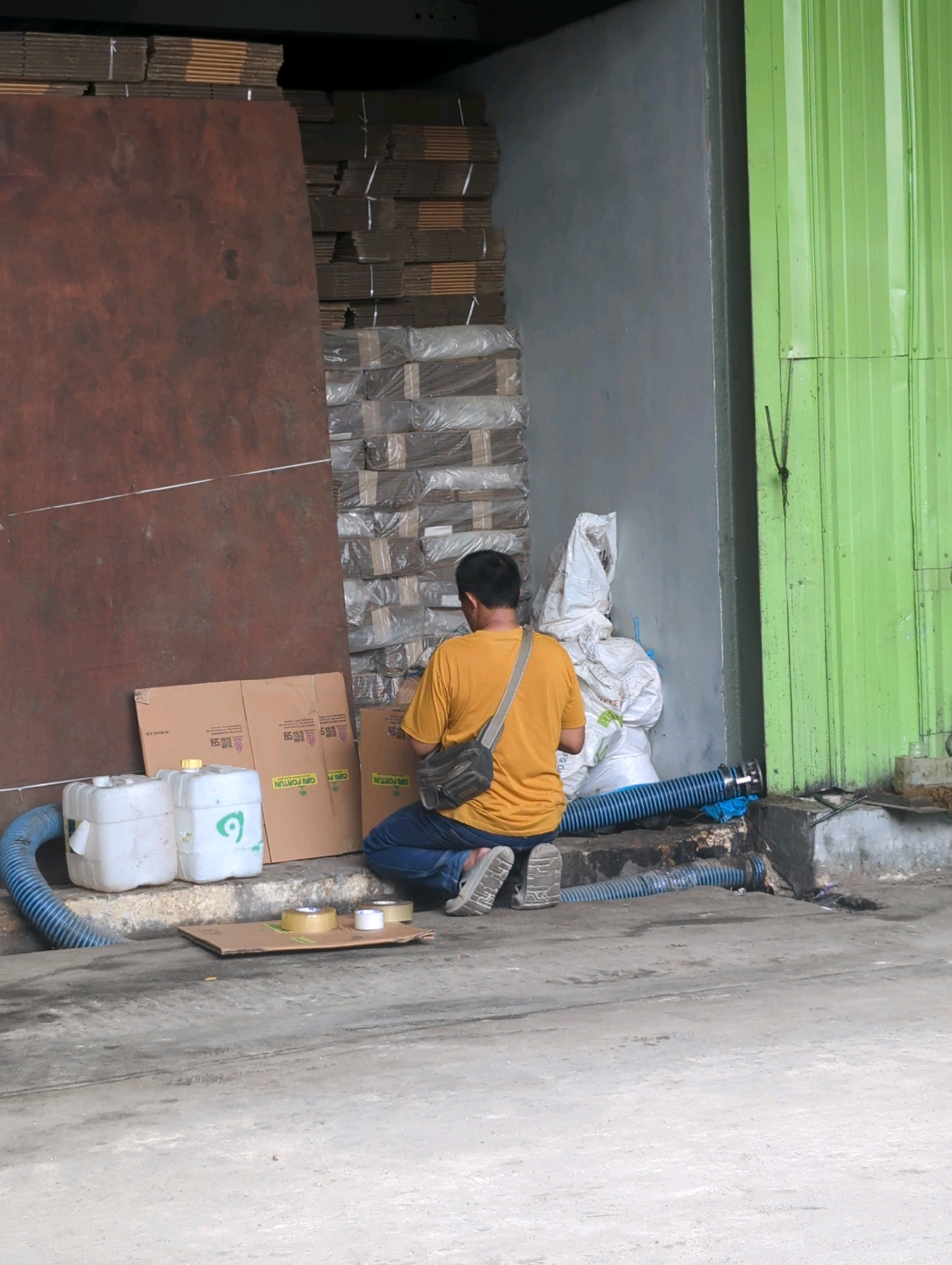 A person kneels in front of stacks of boxes and white sacks next to a grey concrete wall and a vibrant green metal door. The person, wearing a yellow t-shirt, blue jeans, and a dark messenger bag across their chest, is facing away from the camera. In front of them are two white plastic containers, one with a green symbol. To the right of the containers, a large blue corrugated pipe is laid out on the ground, connected to a grey concrete structure. Two rolls of tape sit on a piece of cardboard on the ground. In the background, the wall is covered by stacks of brown cardboard boxes, some wrapped in plastic. The scene appears to be an outdoor industrial or storage area in Batu Belaman, Indonesia.