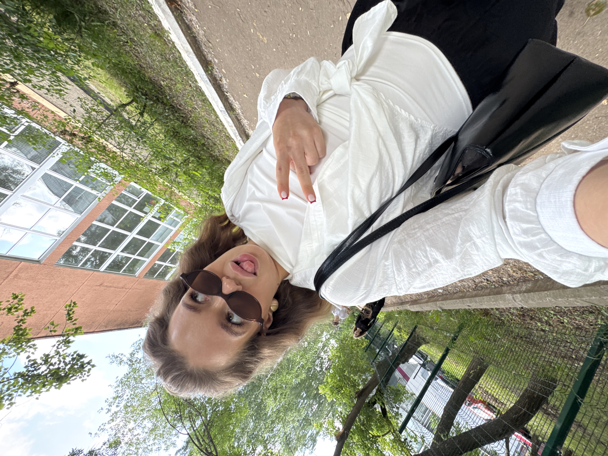 A young woman with brown hair, wearing sunglasses, a white blouse, and black pants, is taking a selfie outdoors. She is sticking her tongue out and making a peace sign with her hand. A black purse hangs from her shoulder. In the background, there is a building with large windows, trees, a fence, and a pathway. The scene appears to be in an urban or suburban setting.