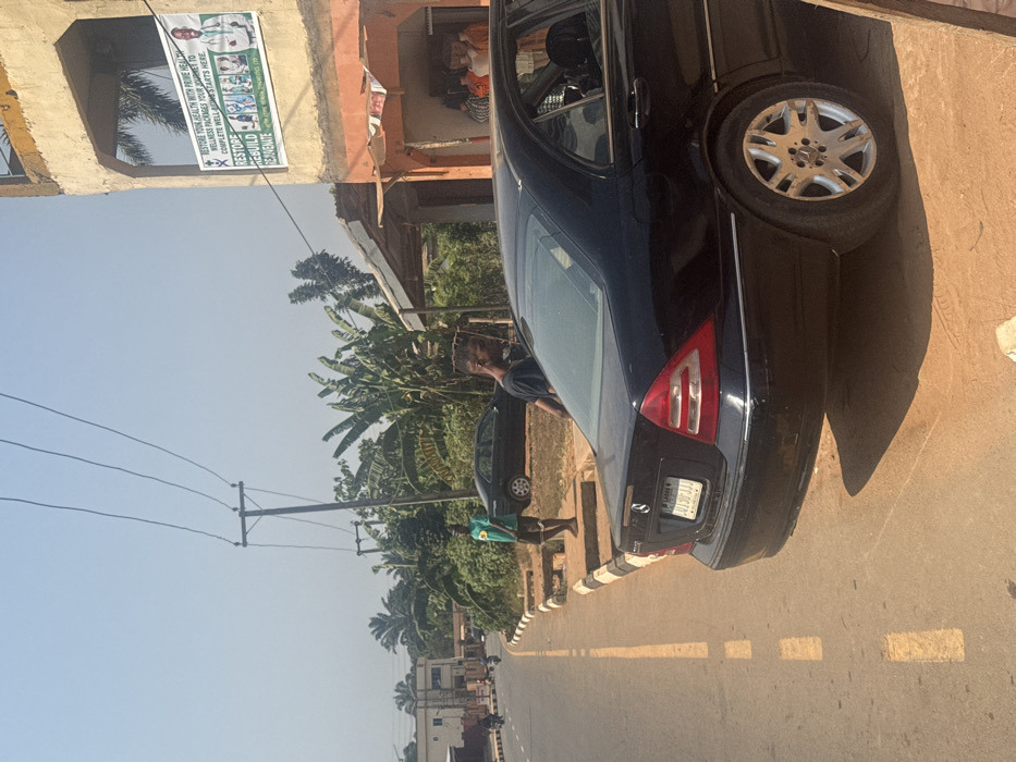 The scene depicts a daylight street view in Aguata, Nigeria, under a clear sky with strong sunlight casting distinct shadows. In the immediate foreground and midground, a black Mercedes-Benz sedan (Lagos license plate "01 JIJ JJJ") is parked off the asphalt road, positioned on a reddish-brown dirt shoulder. A male individual, wearing a dark t-shirt, stands adjacent to the car, partially turned away from the camera. To the left, the road features a solid yellow line on the far edge and dashed yellow markings further inward, bordering a black and white striped curb. A second male individual, wearing a green t-shirt with a yellow emblem and dark shorts, stands barefoot on the raised curb area.
In the midground and background, utility poles with multiple overhead power lines are visible against the sky. Dense tropical vegetation, including large-leafed plants and palm trees, occupies the space behind the parked vehicles. On the right, a two-story concrete building with a weathered facade features an open storefront on the ground floor, revealing several indistinct figures in orange attire inside. The upper floor displays a large banner advertising "Prime Health" wellness packages, with text such as "RESTORE YOUR HEALTH" and "RESTORE REBUILD REJUVENATE," accompanied by images of medical professionals. Further down the road on the left, another dark-colored sedan is parked on a dirt shoulder, partially obscured by foliage, with additional buildings and faint vehicle movements visible in the distance.