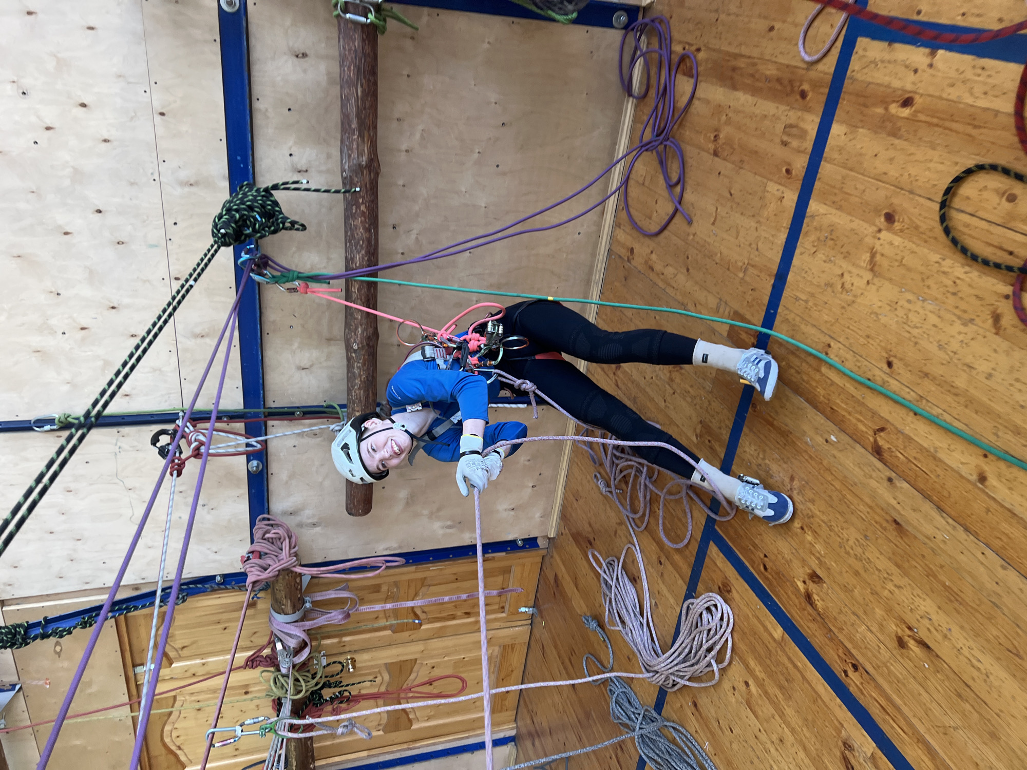 A young woman is suspended on a complex network of ropes and pulleys inside an indoor climbing facility. She is wearing a blue long-sleeved shirt, black leggings, white sneakers, a white helmet, and gloves. The ropes are various colors including green, purple, pink, and grey. She is smiling at the camera. The walls are wooden and there are wooden poles connected to the wall structure. The facility is likely located in Chaikovsky, Russia.