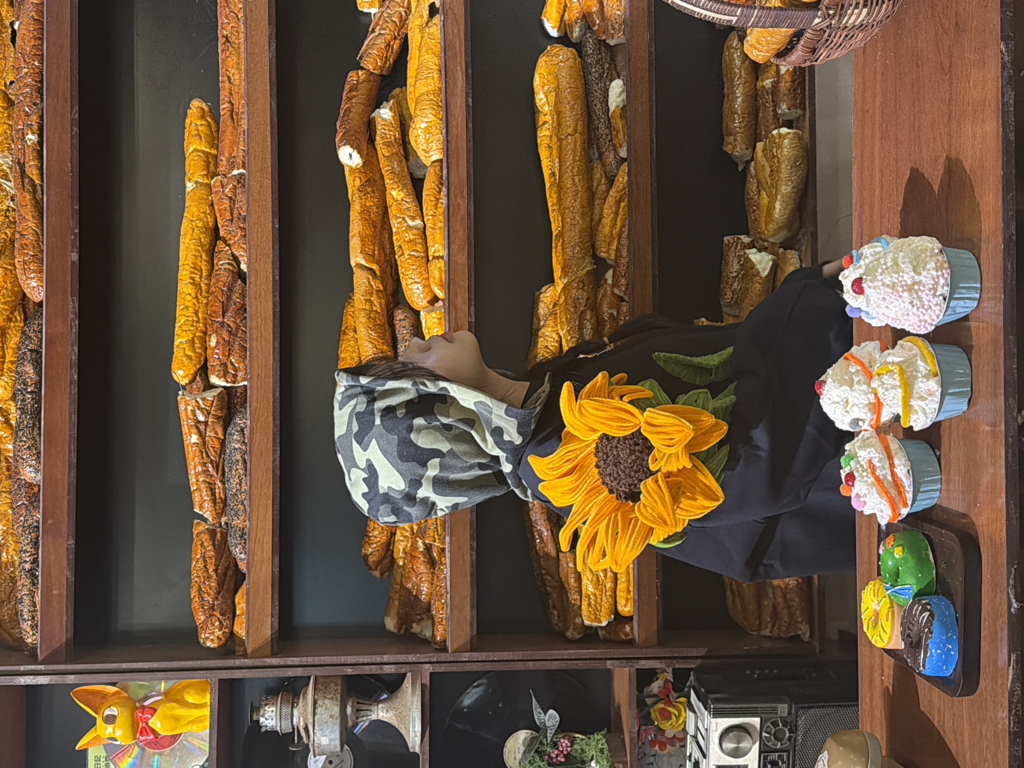 A person wearing a camouflage hat and a black top with a large sunflower design is partially visible in front of a wooden shelf filled with artificial bread loaves. The shelf contains various other items including a yellow rabbit toy, vintage machinery, and floral arrangements. A wooden tray displays artificial cupcakes and donuts. The location is Thuận Hóa Ward, Vietnam.