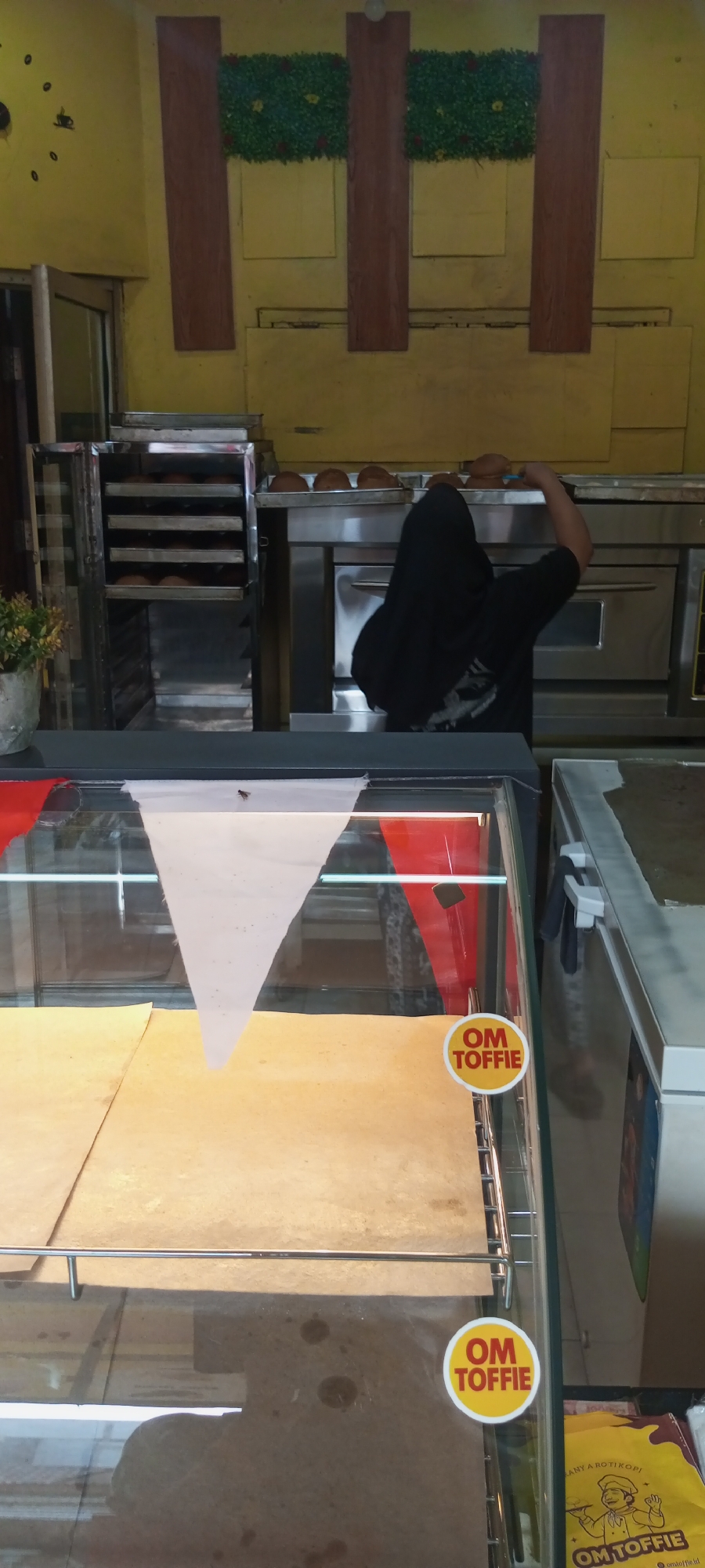 In Setu, Indonesia, a bakery is depicted with a focus on its baking area and display cases. The scene shows an industrial oven with multiple racks containing baked goods, likely bread or pastries. A person, dressed in dark clothing and a headscarf, is partially visible interacting with the oven. In front of the baking area, a glass display case runs horizontally across the foreground. The case contains baked goods on paper-lined trays. Red and white triangular flags are draped inside the display case, and a sticker reading "OM TOFFIE" is visible on the glass. To the right of the display case, a white freezer unit is partially visible, and another "OM TOFFIE" sticker is affixed to its side. The walls in the background are painted yellow and feature decorative elements resembling wooden beams and green foliage. A clock with a minimalist design is mounted on the left wall. A fly is also visible resting on the white triangular flag within the display case.