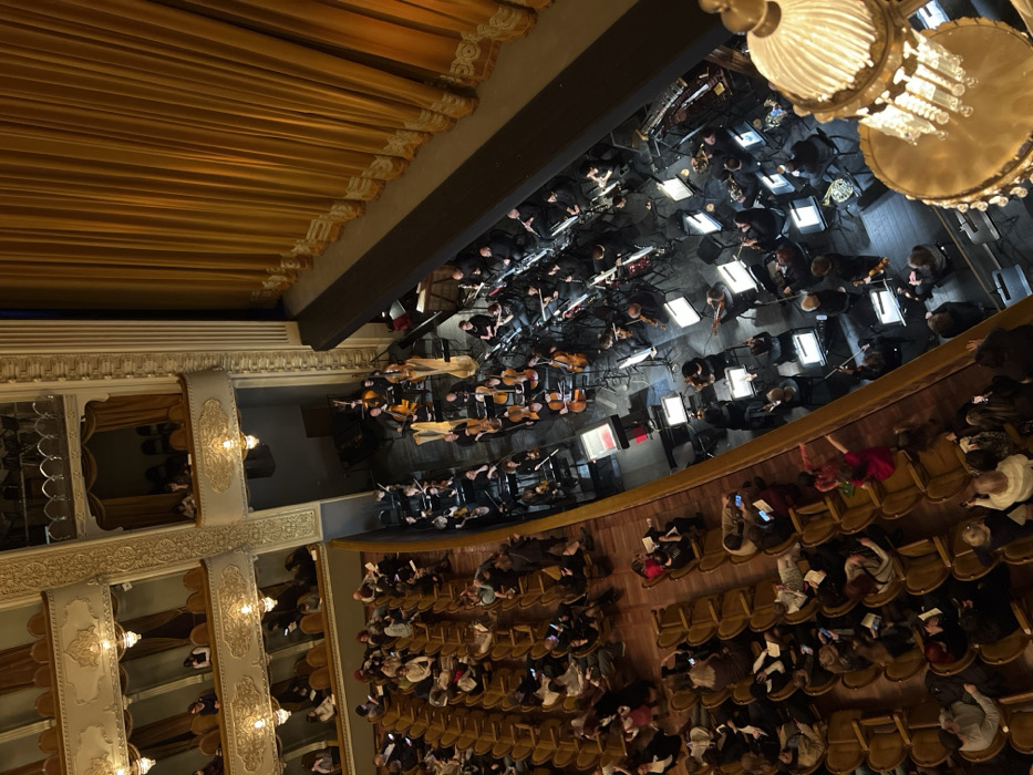 An orchestra performs on a stage viewed from above. The orchestra members are seated and playing instruments. A large chandelier hangs above them. An audience is seated in rows of chairs, many looking at their phones. The interior architecture features decorative molding, balconies, and ornate lighting fixtures, with a golden curtain along the top of the frame.