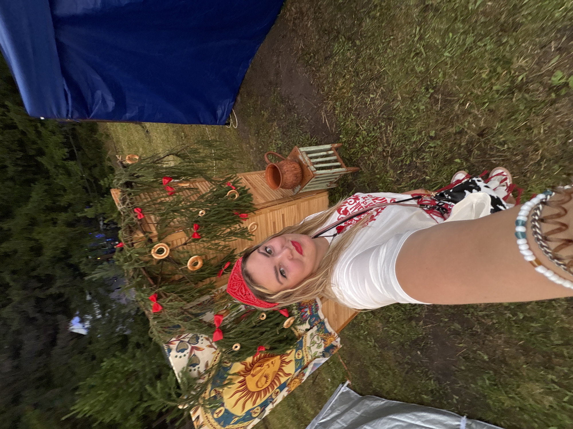 A woman in a red bandana and white t-shirt takes a selfie in front of a wooden structure decorated with greenery, red bows, and round, dry bread rings. To her left is a tapestry with a sun motif. In the background, trees are visible, and to her right is a large blue tent. A small, weathered stool with a clay jug sits near the structure. The scene appears to be outdoors on grassy ground. The location is the village of Polevaya, Russia.