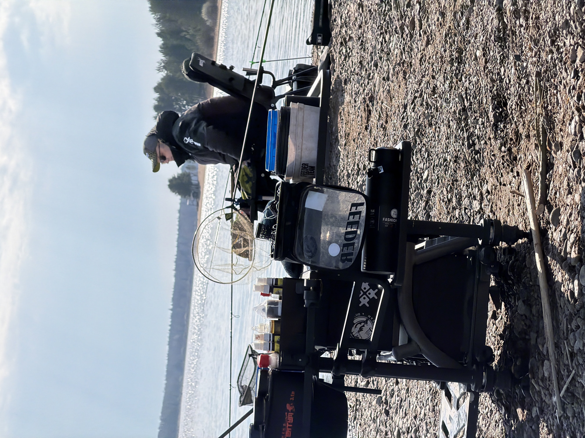 An outdoor scene captures a person engaged in fishing on the pebbled shoreline of a large body of water, likely a lake or reservoir near Chaykovsky, Russia. The image is originally rotated 90 degrees clockwise but depicts a landscape view. A person, dressed in a dark jacket, a baseball cap, and glasses, is positioned on the left, appearing focused on their fishing setup as they look towards the water.
In the foreground and midground, an elaborate fishing station is set up. This includes a robust black fishing chair or platform, supporting multiple fishing rods with reels, a large circular landing net, and various tackle and bait containers. A black thermos or bottle is also visible. The ground beneath the station is covered in small, light-colored pebbles, characteristic of a shoreline.
The background features a wide expanse of calm, glistening water, reflecting the bright sunlight. A distant treeline borders the far side of the water under a clear, bright blue sky with sparse clouds. The lighting indicates it is late morning or early afternoon, with distinct shadows cast by the equipment.
Several pieces of text are visible on the fishing gear: "FEEDER" and "FASHION (316)" are seen on a clear pouch and a black bottle, respectively, while "FEEDER TEAM" is on a grey container, suggesting a focus on feeder fishing. "VENTUM 3.1in" is visible on a black bag. A sticker with "DB MVPS" and a graphic is affixed to the fishing station, and "DG" is visible on a rod bag. The overall atmosphere is peaceful and dedicated to the activity of fishing.