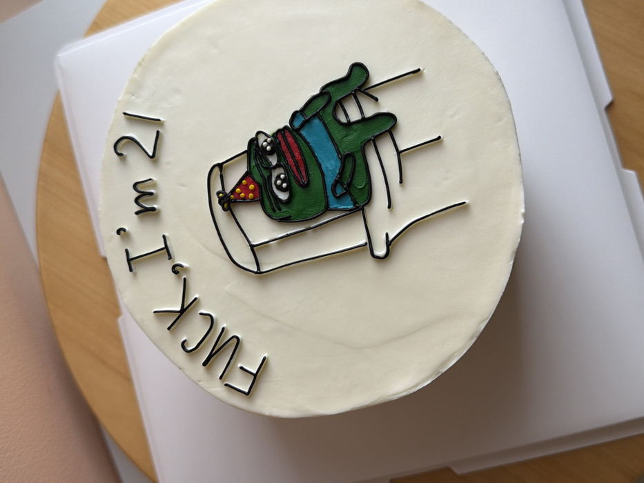 A round cake with off-white frosting is depicted resting on a white cake board, which is partially visible on a light brown wooden surface. The cake's top features black piped frosting text stating "FUCK, I'm 2/". Below the text, a cartoon character resembling Pepe the Frog is rendered in green, wearing a blue shirt and a red party hat with yellow polka dots. The frog has a red open mouth and prominent eyes, and is depicted sitting on a white toilet, outlined in black. The background includes the light brown wooden surface and an out-of-focus light beige or white surface.