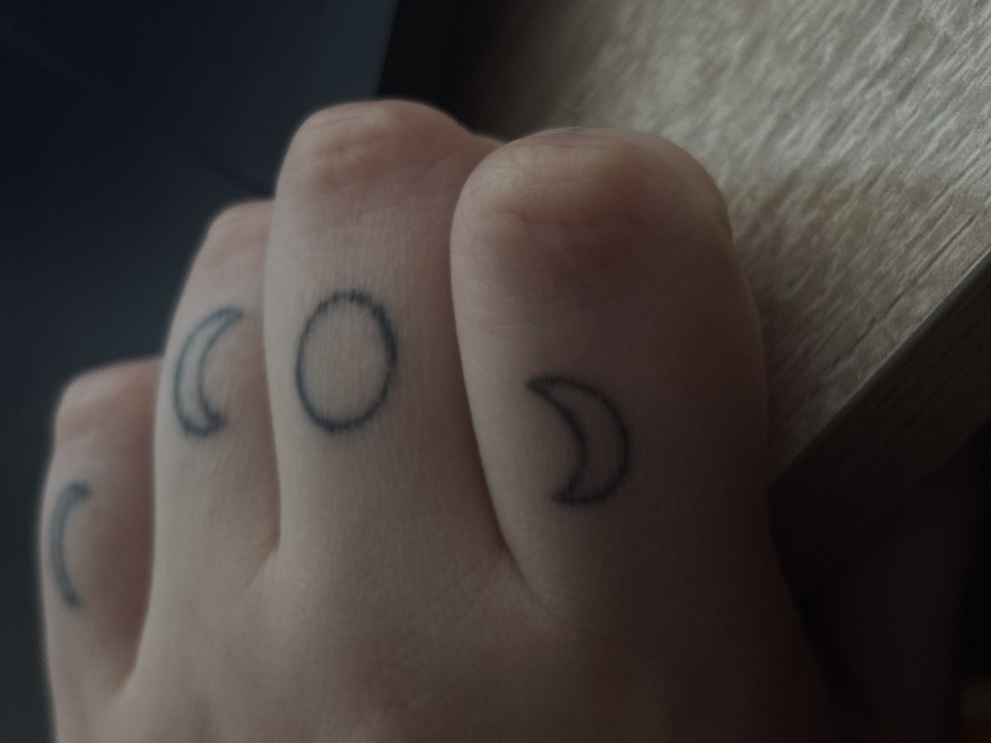 A close-up, slightly out-of-focus shot shows a hand with several small, thin-lined tattoos on the fingers. The tattoos appear to be phases of the moon: a crescent moon on the pinky finger, a full circle on the ring finger, and another crescent moon on the middle finger. There is a partial crescent moon tattoo visible on the index finger as well. The hand is resting on a light-colored wooden surface, possibly a desk. The background is dark and indistinct, suggesting an indoor environment. The lighting is dim, casting a soft glow on the hand. The image is taken from a low angle, looking up at the hand. There is no identifiable location information in the image that would place it in Saint Petersburg, Russia.