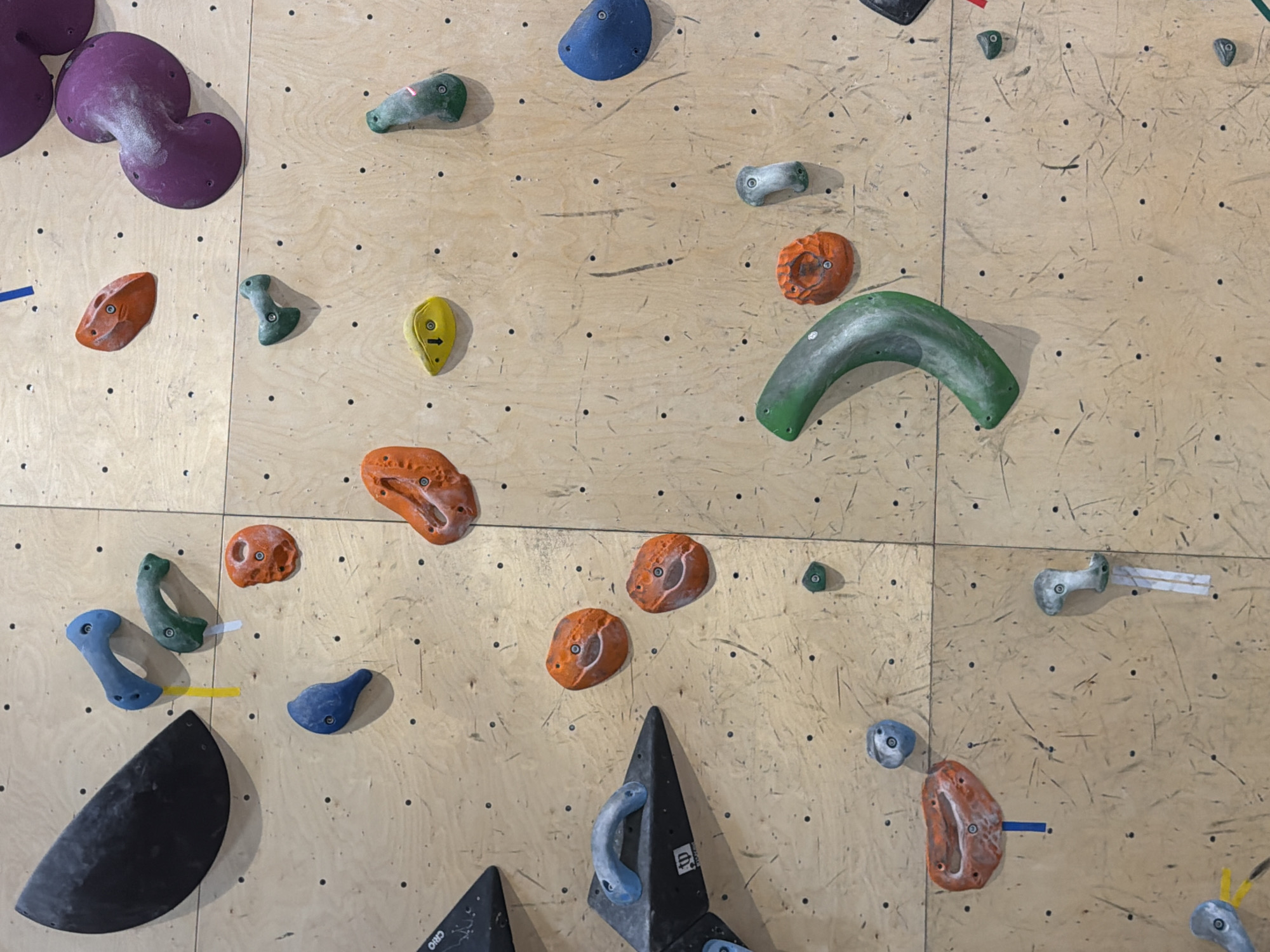 The image depicts a climbing wall, likely indoors, constructed from plywood panels and featuring a variety of colorful climbing holds of different shapes and sizes. The holds are arranged seemingly randomly across the surface. There are no people visible in the scene. There are no explicit clues to confirm that the image was taken in Yerevan, Armenia.